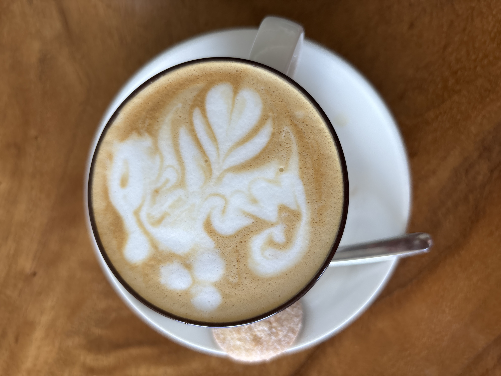 An overhead shot reveals a cup of coffee, seemingly a latte, adorned with latte art, resting on a white saucer. A small, round cookie sits on the saucer's edge beneath the cup. A silver spoon is also positioned on the saucer. The scene is set on a wooden surface. There is no specific information in the image to confirm that the location is in Kuta Selatan, Indonesia.