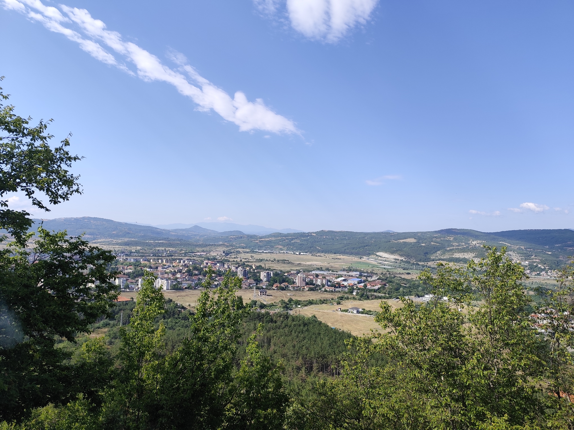 A wide-angle, slightly high-angle shot captures a landscape view of Momchilgrad, Bulgaria. The foreground is dominated by lush green trees, with their branches and leaves obscuring parts of the view. Beyond the trees, a town is nestled in a valley. Buildings of various heights and colors, including apartments, houses, and commercial structures, are visible. Some buildings are white, while others have red roofs or are constructed from brick. Open fields, some dry and golden, others green, surround the town, interspersed with more forested areas and rolling hills in the distance. The sky is a clear blue, with a few wispy white clouds streaking across it. The overall impression is a serene, sunlit day overlooking a Bulgarian town.