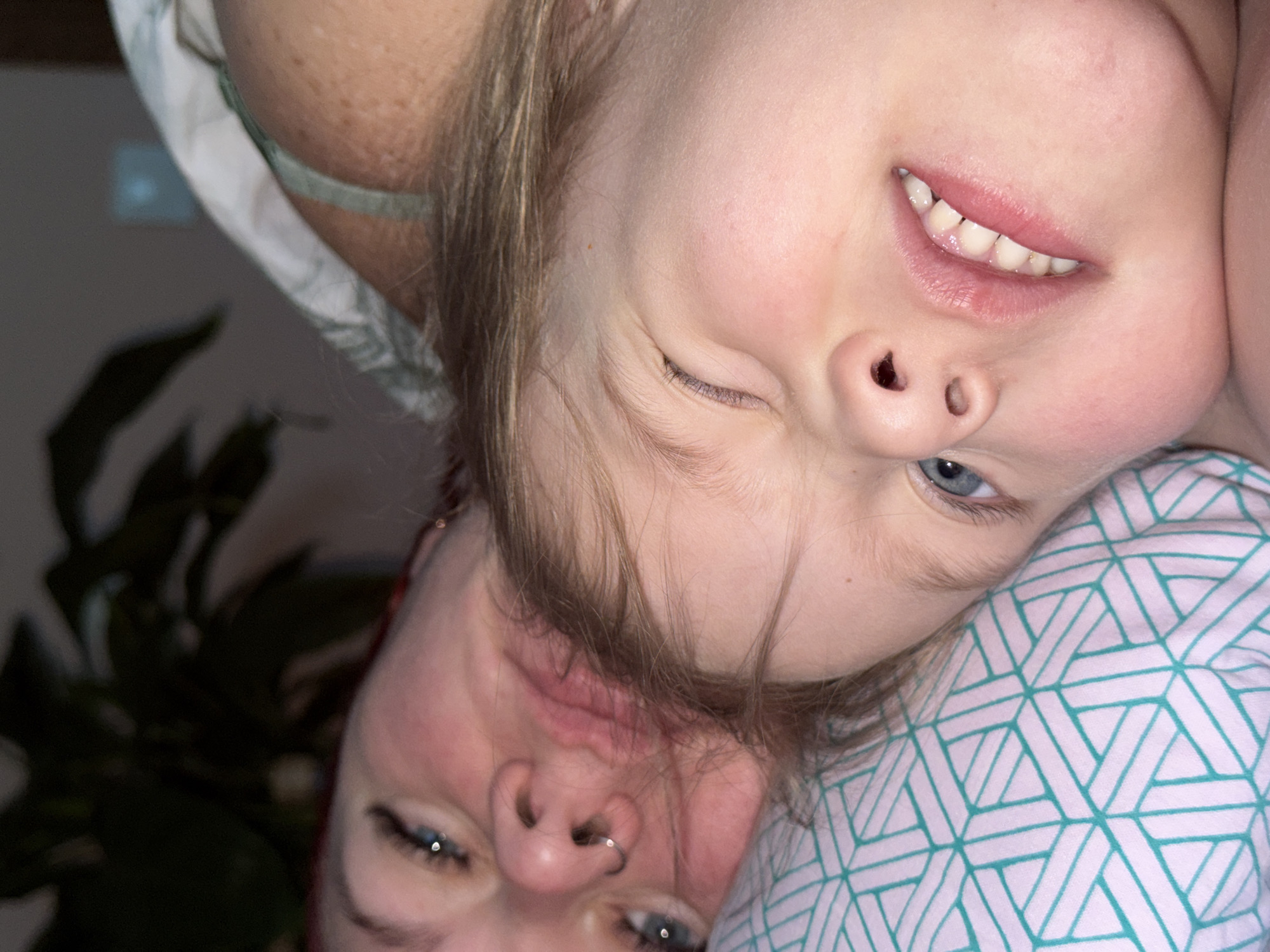 A close-up, upside-down shot features two individuals. The first is a young child, possibly a girl, who is winking and smiling slightly. Her head rests on a pillow with a repeating geometric pattern of blue-green lines on a white background. The second individual, an adult, is positioned below the child, also upside down. The adult's eyes are open and focused, and a nose ring is visible in the left nostril. To the left of the adult's face, a blurred green plant provides a backdrop. The location is unknown. The city of Chaikovsky, Russia is also unknown.
