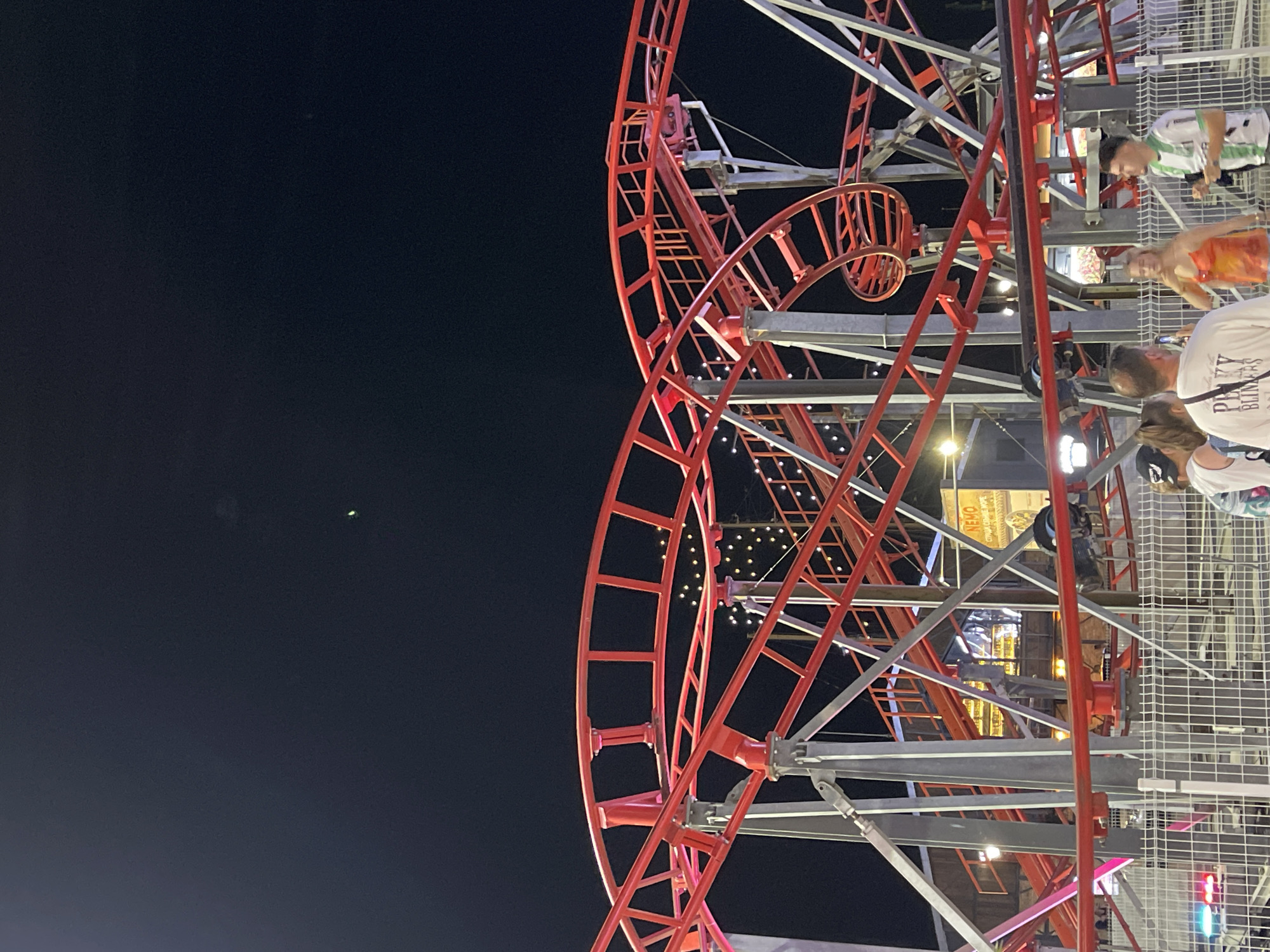 This image captures a portion of a red roller coaster at night in Varna, Bulgaria. The roller coaster's track and support structure are prominently featured, with decorative lights visible in the background. In the foreground, four individuals are standing near a fence, appearing to observe the roller coaster. The scene is dimly lit by the ambient lighting of the amusement park.