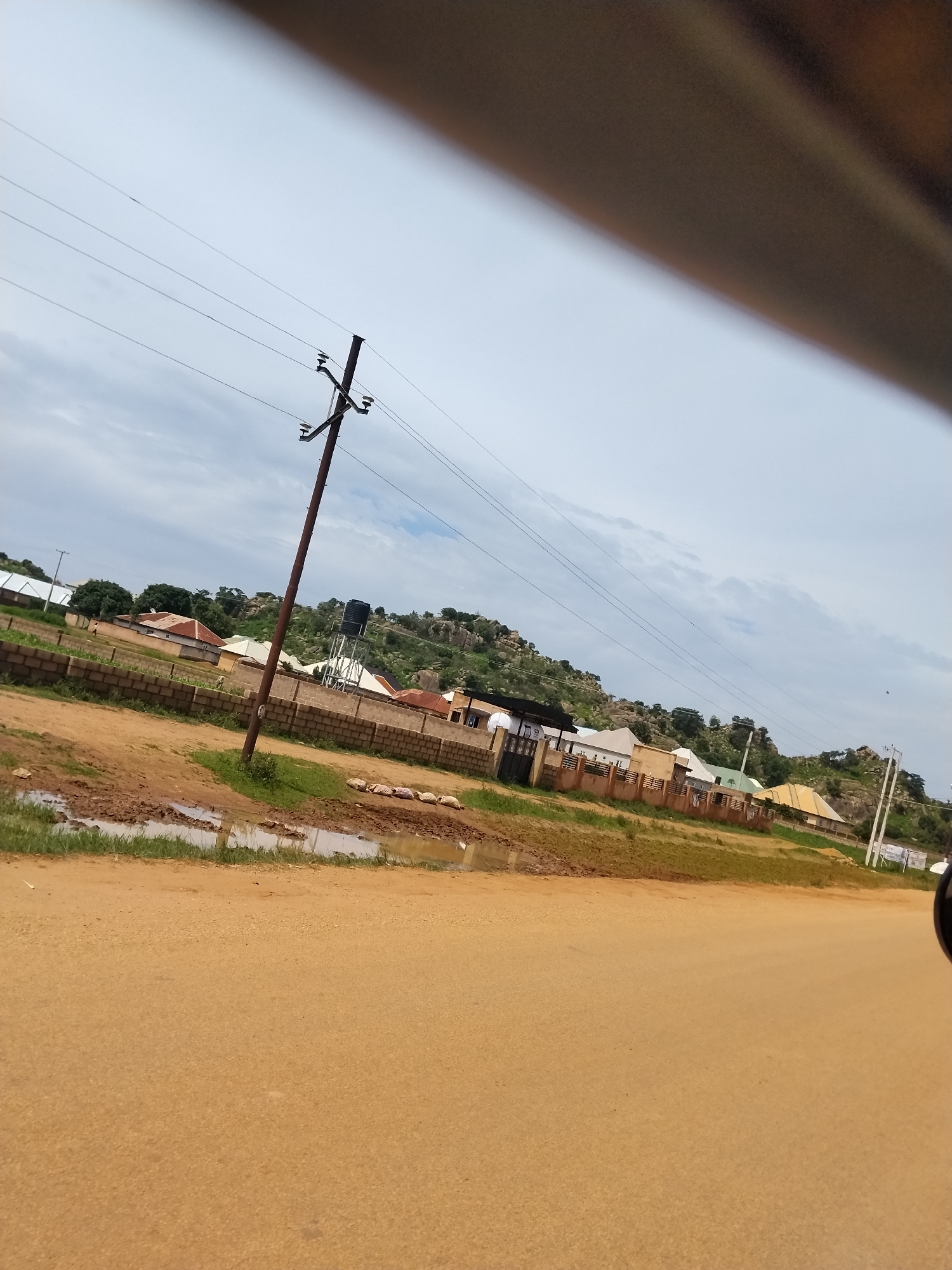 The image shows a scene in Nabatum, Nigeria, featuring a dirt road in the foreground with puddles of muddy water and some sandbags. A tall, wooden utility pole with wires runs diagonally across the frame from the middle left to the upper left. In the middle ground, a low, brick wall borders a grassy area with several houses and buildings, some with corrugated iron roofs and others with lighter-colored roofing materials. A water tower is visible behind the houses on the right side of the image. The background consists of a rocky, tree-covered hillside under a cloudy sky. The overall environment appears to be a rural or semi-urban residential area.
