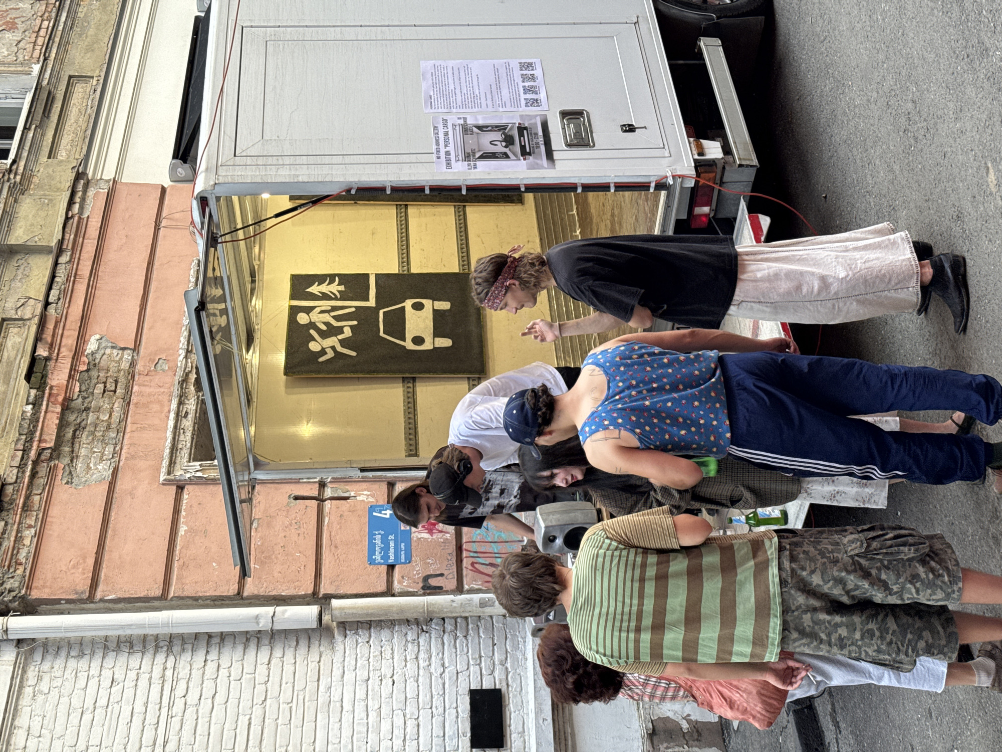 In Tbilisi, Georgia, a group of young adults is gathered around an open-back truck, which appears to be serving as a mobile exhibition space or stall. The truck's interior is illuminated, displaying a large black and white graphic with stylized figures and a car. Posters are attached to the side of the truck, one of which is a flyer for an exhibition titled "PERSONAL CARGO" and lists DJ performances. A street sign above the group indicates "Vashlovani St." The individuals are dressed in casual attire. Some are looking at a small speaker system on a table, suggesting music is being played or set up. One person is holding a green bottle. The buildings in the background show signs of age, with peeling paint and weathered brickwork. The scene takes place on a paved street.