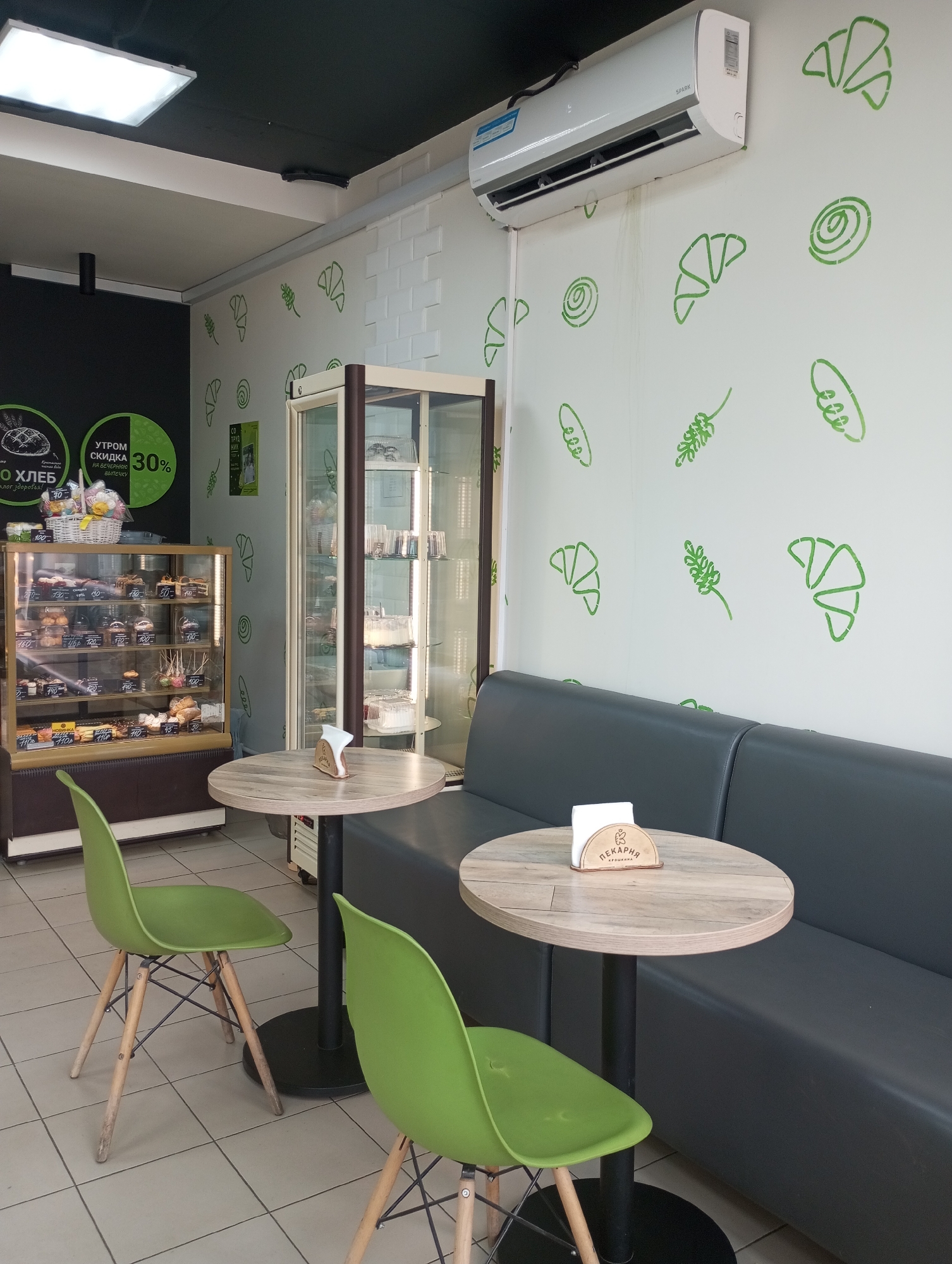 This is an interior shot of a bakery, likely located in Izhevsk, Russia, based on the Cyrillic text on the signage. The bakery has a modern, minimalist design. There is a refrigerated display case filled with pastries and cakes on the left side of the frame. A second display case with pastries is situated toward the back. Two small round tables with green plastic chairs are positioned in the foreground. A gray upholstered bench seat runs along the right wall, which is decorated with stenciled images of bread and croissants. An air conditioning unit is mounted high on the wall. The flooring is tiled.