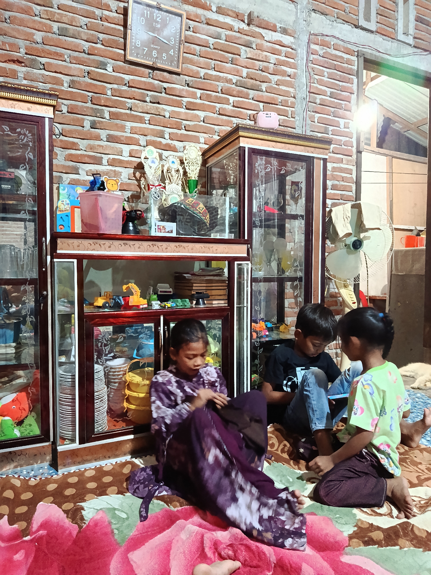 A group of four children are gathered on the floor in a room with a brick wall. Two glass display cabinets filled with various items are prominent features of the room, one on the left and another in the center. The cabinet on the left appears to contain plates and various containers, while the central cabinet displays toys, including toy vehicles, and other decorative items. On top of the central cabinet, there are trophies and what looks like a pink portable speaker. A wall clock hangs on the brick wall, indicating the time.
Three of the children are actively engaged in an activity on the floor. A girl in a purple patterned dress is seated, appearing to be looking at something in her lap. Next to her, two children, a boy in a black t-shirt and jeans, and a girl in a green t-shirt, are looking down at a device, possibly a tablet, held between them. Their focused expressions suggest an engaging activity. Another child's leg is visible in the foreground, suggesting they are also part of the group, though their face is not shown. A fan is visible in the background. The overall atmosphere appears to be one of casual domesticity, possibly in the afternoon, given the lighting. The room seems to be a living space, likely in Indonesia, as indicated by the location context.