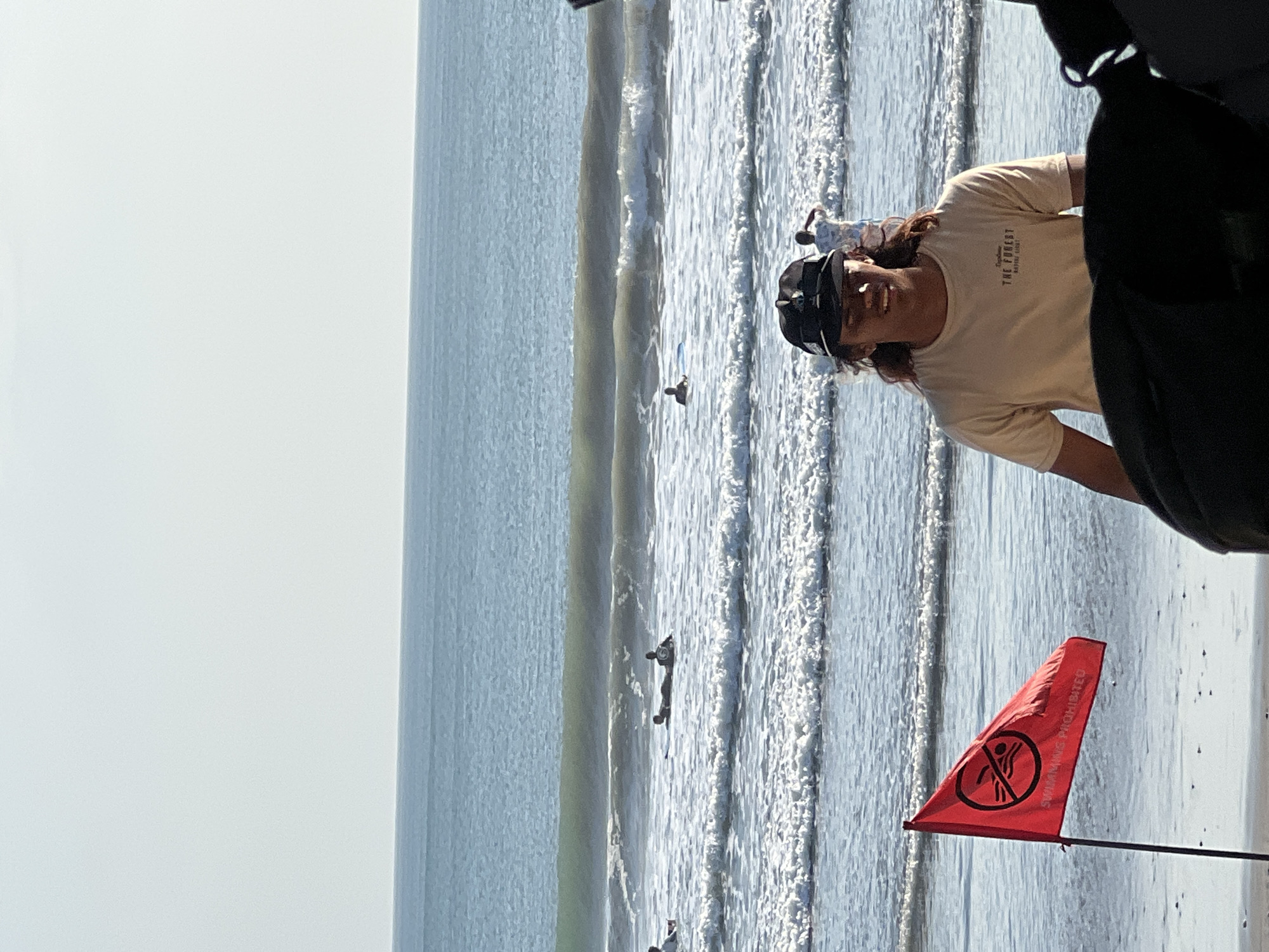 This is a photograph taken at Kuta Beach in Kuta, Indonesia. In the foreground, a young man wearing a tan t-shirt and a black baseball cap stands facing the camera, with his long, dark hair flowing. To his left, a red flag on a pole, bearing a circle with a diagonal line through it over a swimmer icon and the text "SWIMMING PROHIBITED," is planted in the sand. In the background, the ocean has moderate waves, and several individuals are visible in the water, some on surfboards. The sky is clear and bright, suggesting a sunny day.
