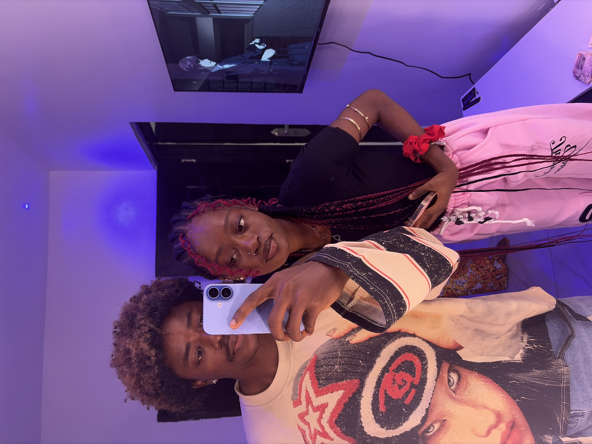 Two individuals are captured in what appears to be a mirror selfie. One person has afro-textured hair, wearing a patterned t-shirt with a large graphic image of a woman's face. The other individual has dark hair styled in braids with red highlights, wearing a black top and pink sweatpants. The person with the phone is holding a blue-colored smartphone up to take the selfie.
The setting includes a television displaying an anime scene, a shelf, and purple-blue lighting in the room. The location is Igboefon, Nigeria.