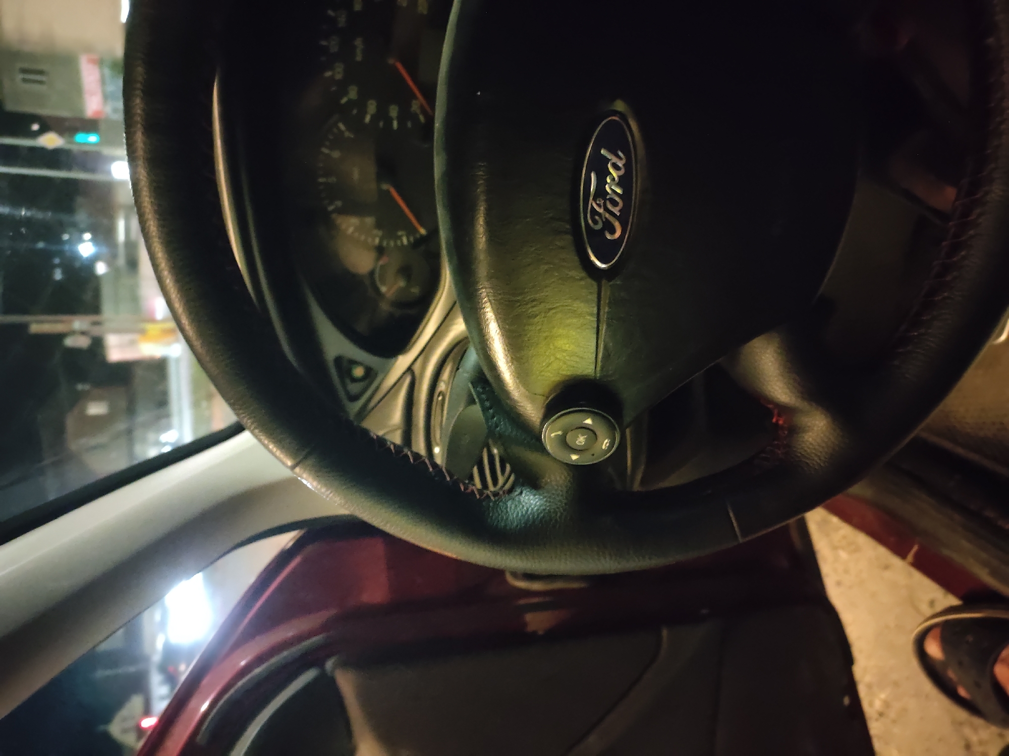 The image depicts the interior of a car at night. A black leather steering wheel with red stitching is prominent in the foreground, featuring a Ford logo in the center. To the left, the red interior door panel and a portion of the car's pillar are visible. The dashboard, including the speedometer, is partially visible behind the steering wheel. Outside the car, streetlights and buildings are faintly discernible, suggesting an urban or suburban environment at night. The lower left corner of the image shows a person's foot wearing a black sandal. There is no specific indication that the location is Leninkent, Russia.