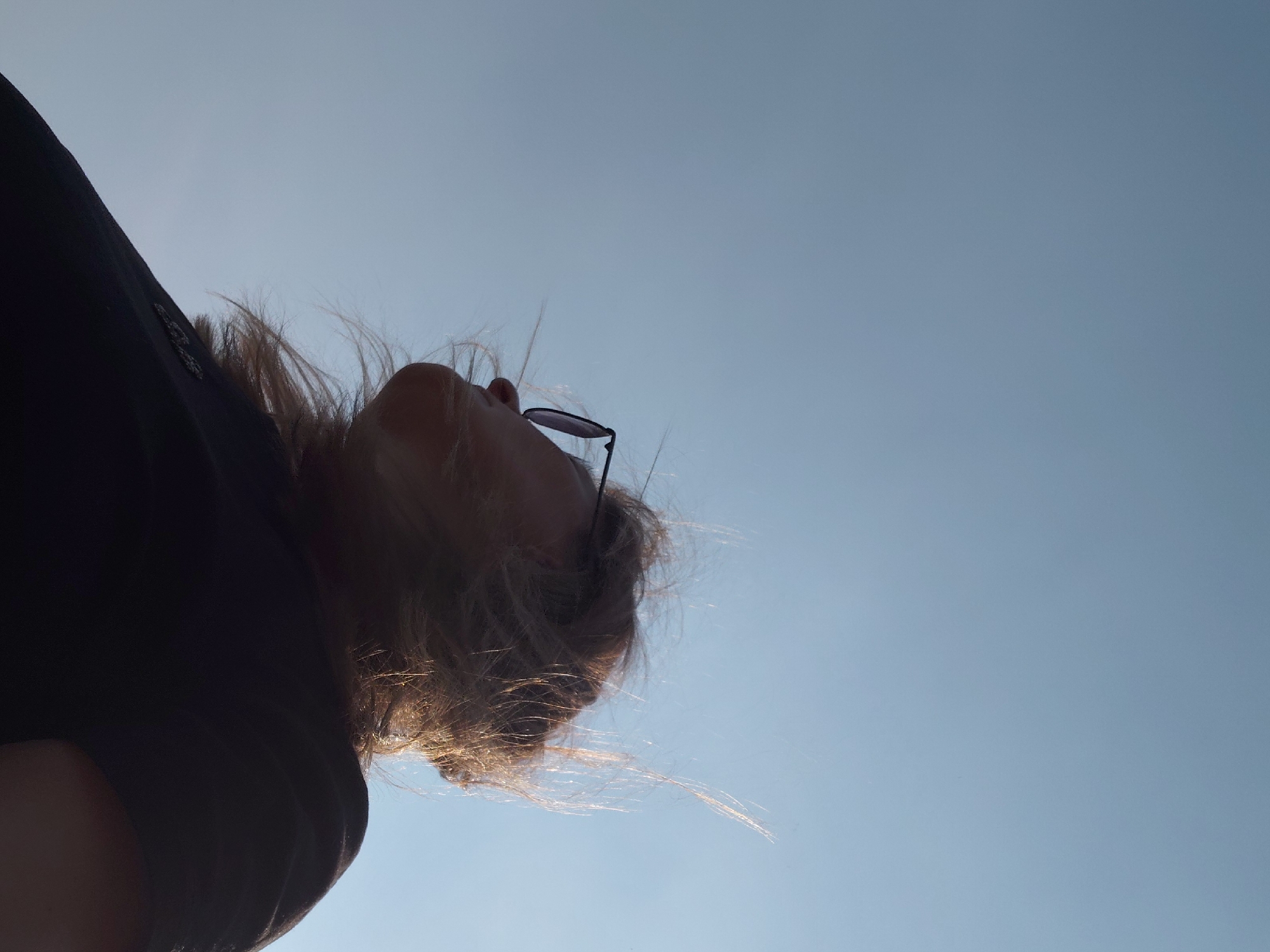 A low-angle, full-body outdoor shot shows a person looking up at a clear, pale blue sky. The person is wearing a dark t-shirt and sunglasses. Their hair is blowing in the wind, with some strands illuminated by sunlight. The image appears to be taken during the daytime. The specific location in Novosibirsk, Russia, cannot be determined from the visual information provided.