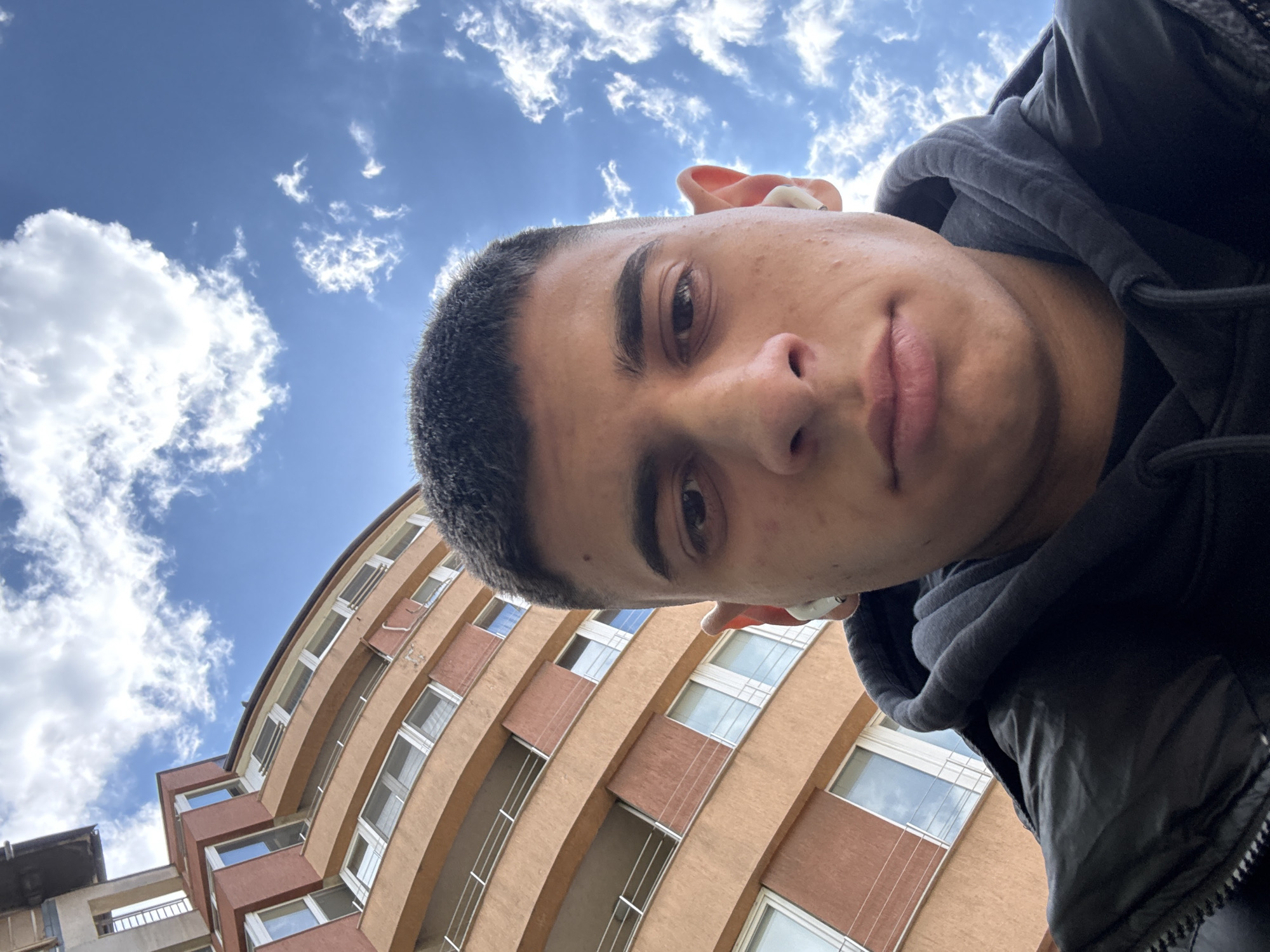 A low-angle selfie photograph shows a young man with short dark hair, fair skin, and brown eyes. He wears a dark hooded jacket. A white wireless earbud is visible in his ear. Behind the man, a light-brown, multi-story apartment building with multiple windows is visible. The sky is blue with scattered white clouds. Based on architectural style, the location is likely in the city of Sofia, Bulgaria.