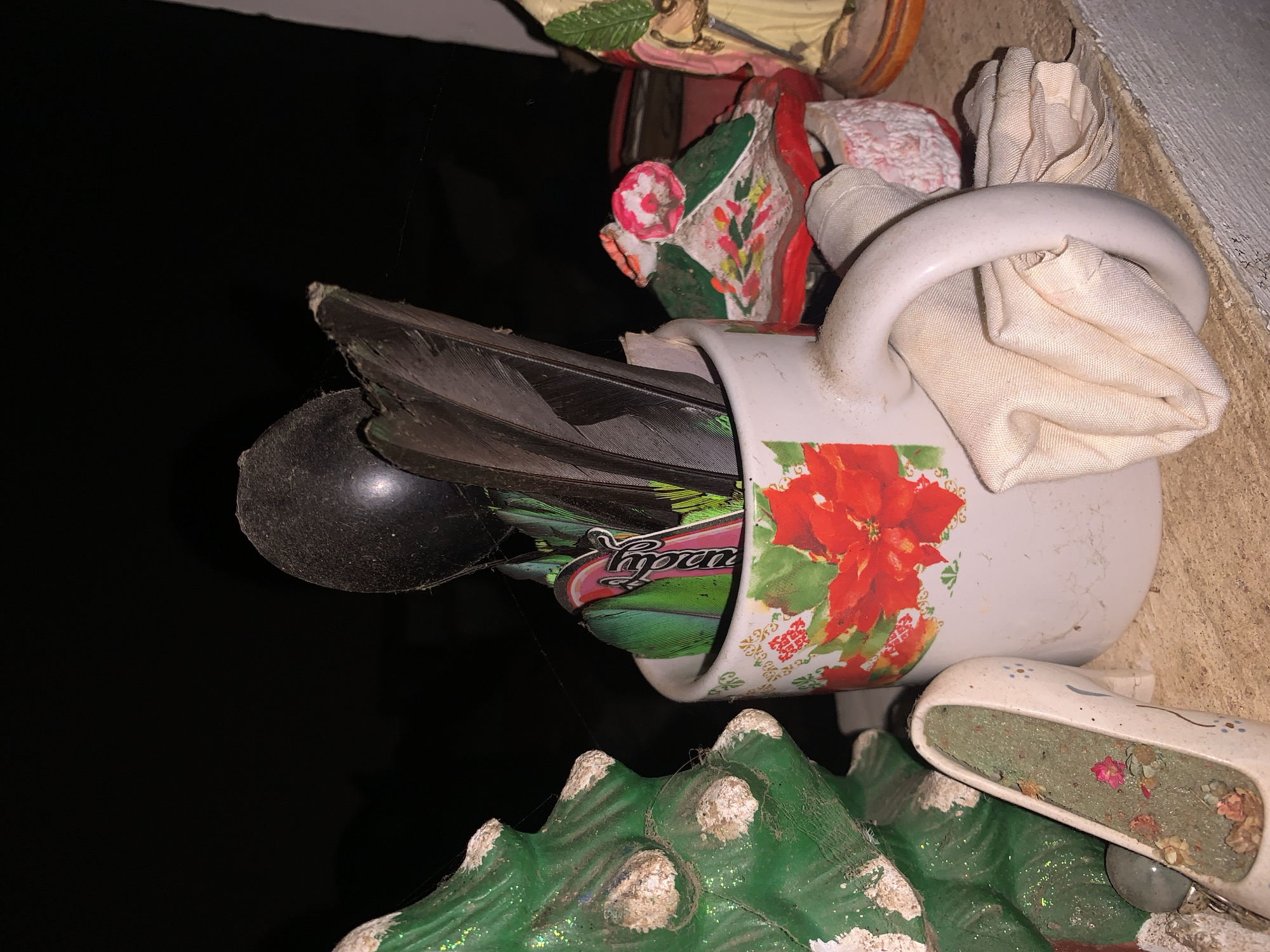 This close-up image, horizontally oriented, depicts a collection of decorative items on a surface, likely a shelf or ledge, set against a dark background, suggesting nighttime or a deeply shaded environment.
The central piece is a white ceramic mug or small pitcher adorned with festive red poinsettias and green foliage. Inside the mug are several dark, iridescent feathers, possibly from a bird, and a smooth black spherical object. A partially visible label inside the mug reads "Yogurt" in a stylized font. Light-colored, crumpled fabric is wrapped around the mug's handle and tucked into its opening.
Surrounding the mug are other decorative elements. Above it are parts of painted ceramic or plaster figurines, one featuring green leaves and a pink pattern, and another showing part of a human figure. Below the mug is a green, textured object resembling stylized mountains or hills, tipped with white, possibly glitter, which could be part of a nativity scene or a landscape decoration. Adjacent to this is a small white shoe-shaped ornament, possibly a clog, filled with dried flowers or potpourri in various shades of green, pink, and brown.
Thin strands of spiderweb are visible clinging to the objects and extending into the dark background, hinting at an aged display. The items collectively suggest a decorative arrangement, possibly for a holiday or personal collection, in a domestic or outdoor setting in Santa Ana, Venezuela. No specific activity is occurring; the image captures a static arrangement.