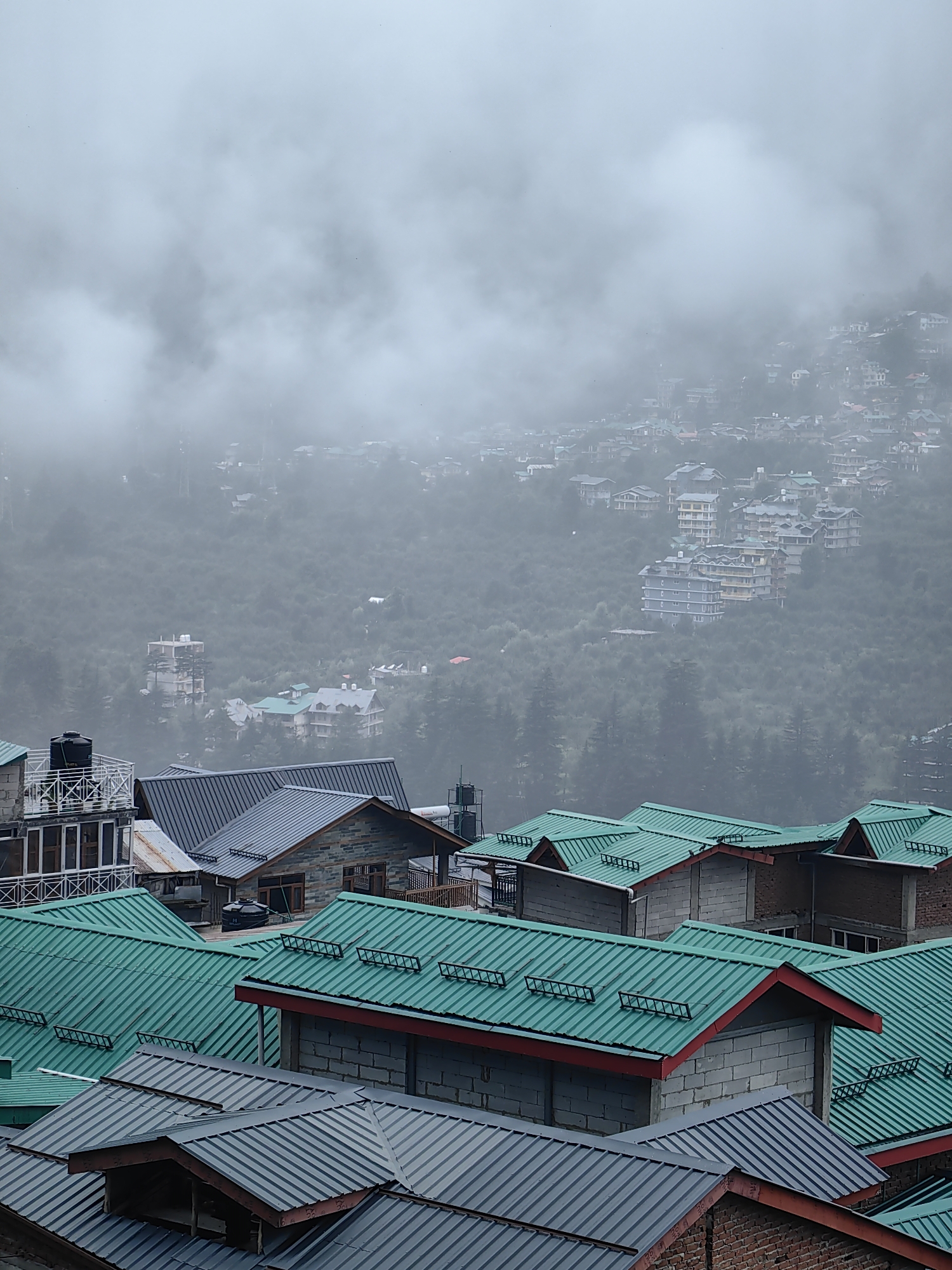 The image depicts a dense residential area nestled on a foggy hillside, likely in Vashisht, India, given the architectural style and terrain. The foreground is dominated by the rooftops of several multi-story buildings. These rooftops are primarily constructed of corrugated metal, with prominent green and gray sections. Many of these roofs feature a zigzag pattern of ridges and valleys, and some are accented with red trim. Small, ladder-like structures are visible on several of the green roofs, possibly for snow or water runoff.
In the midground, more buildings are visible, interspersed with dense green foliage, suggesting a forested hillside. These structures are varied in size and design, with some appearing to be hotels or larger complexes, characterized by multiple stories and balconies. The structures in the midground are partially obscured by the persistent fog, which creates a soft, diffused light and lends an ethereal quality to the scene.
The background is a hazy panorama of a sprawling town or village extending up the mountainside. Numerous buildings, varying in height and color, are visible but are heavily softened by the fog, making individual details difficult to discern. The overall impression is of a community built on a steep incline, with architecture adapted to the mountainous environment. The weather appears overcast and possibly damp, contributing to the pervasive mist. No people or active human interactions are discernible in the image.