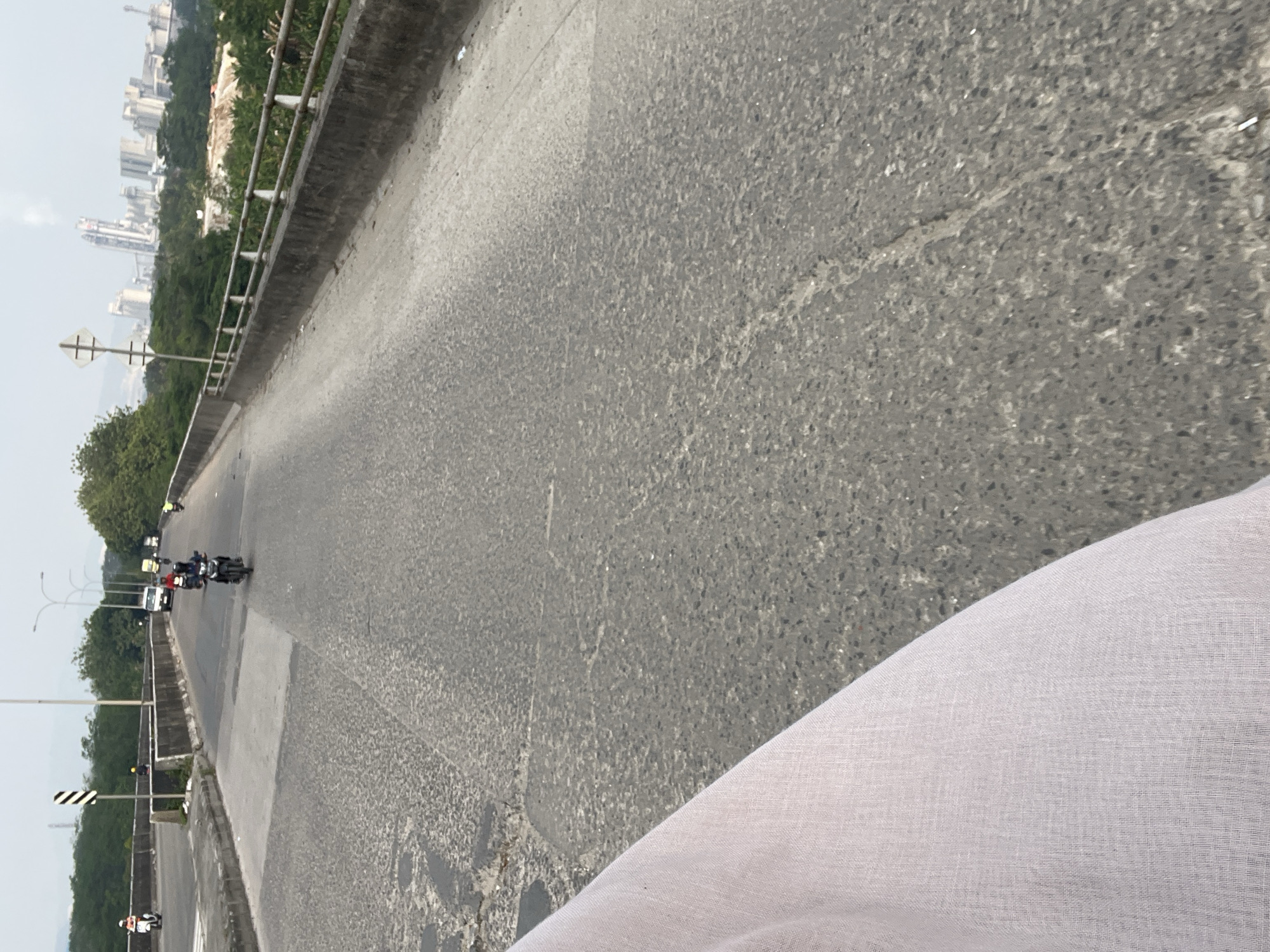The image shows a wide, cracked asphalt road, likely a bridge or overpass, in Gunung Putri, Indonesia. Several motorcycles are traveling on the road, with riders wearing helmets. In the background, a cityscape with tall buildings and industrial structures can be seen, with a slightly hazy sky overhead. Green trees line the sides of the road and the surrounding landscape. A white fabric, possibly clothing, is visible in the lower left corner of the frame. The scene appears to be a typical day with traffic on a major road in an urban or semi-urban environment.