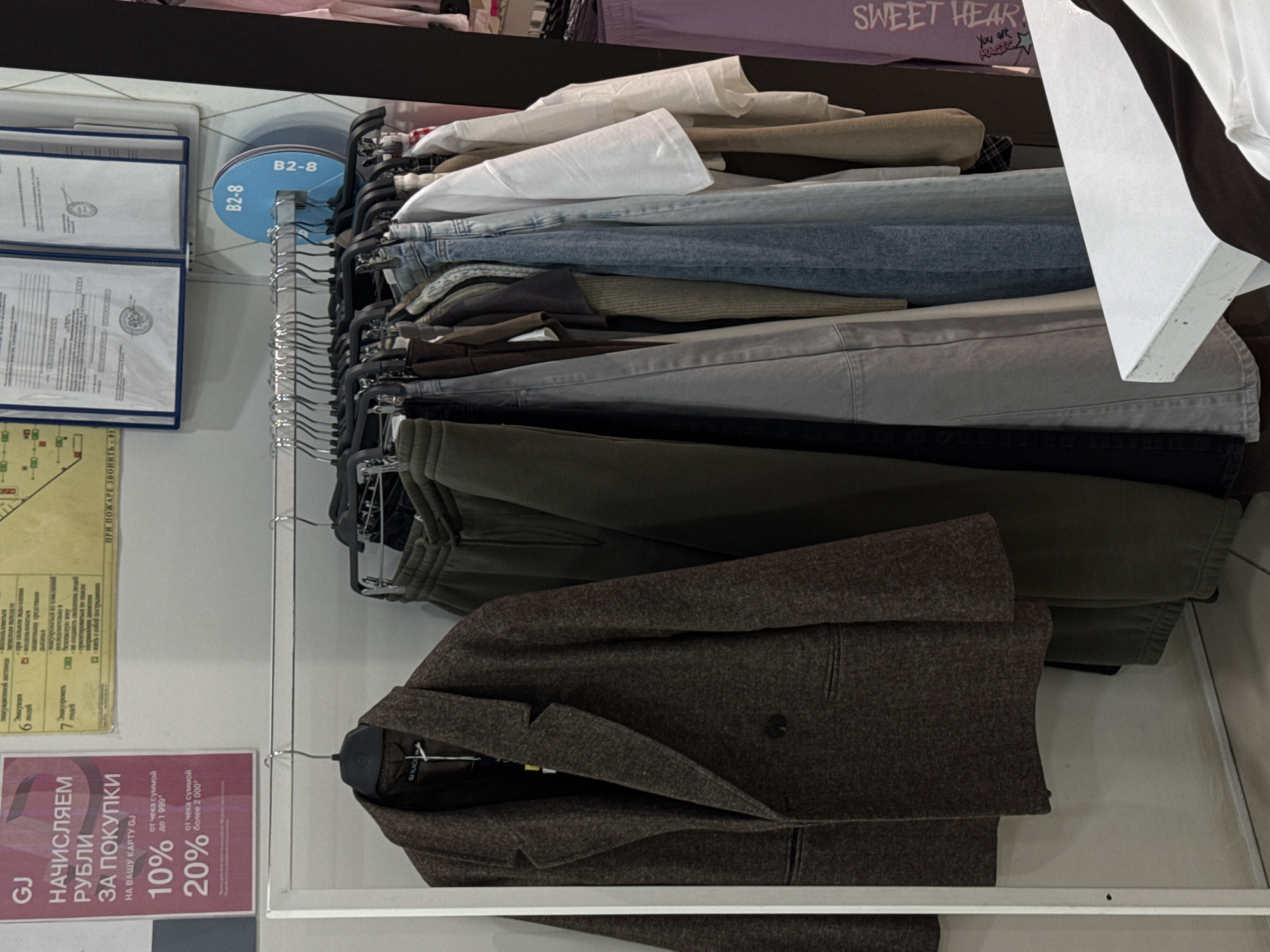 The image captures an interior view of a clothing store, showcasing racks of garments. In the foreground, a dark brown or charcoal-colored tailored jacket or blazer, made from a textured fabric, hangs on a black hanger. Behind it, a clothing display holds a variety of folded and hung items including white and light beige tops, light blue denim jeans, light grey pants, darker trousers, and olive green sweatpants. Above these, more folded clothes are visible on shelves, including a purple garment inscribed with "SWEET HEAR" and "You are MAGIC."
To the left, several framed documents are displayed on a light-colored wall. Below them, a prominent pink and red poster in Russian advertises a loyalty program for "GJ," stating "НАЧИСЛЯЕМ РУБЛИ ЗА ПОКУПКИ НА ВАШУ КАРТУ GJ" (We accrue RUBLES for purchases on your GJ card), with offers of "10% от чека суммой до 1999" (10% from checks up to 1999 rubles) and "20% от чека суммой более 2000" (20% from checks over 2000 rubles). Adjacent to this, a partial poster gives emergency instructions in Russian, including "ПРИ ПОЖАРЕ ЗВОНИТЬ-01" (In case of fire, call 01) and notes about evacuation. A blue circular sign with "B2-8" inscribed, attached to a vertical pole, marks a section of the store. The setting is an organized retail space, with no people visible. The visible text confirms the location context of Perm, Russia.