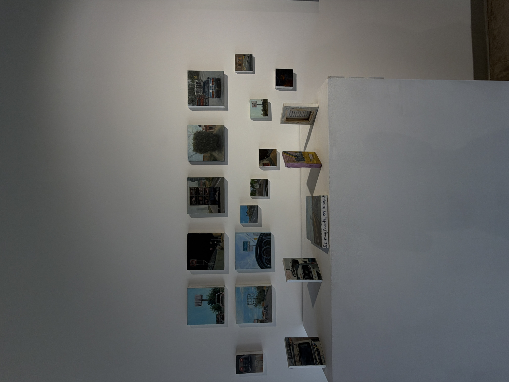 The image shows a wall in an art gallery in the city of Unknown, Unknown. A series of small, square paintings are arranged on the wall. The paintings depict various street scenes, including cars, buses, trees, and street signs. The paintings are mounted on a white wall and are displayed in a seemingly random pattern. Some of the paintings are displayed on a small white shelf that protrudes from the wall. One painting is a book open to a page that has text written in an unknown language. The environment appears to be well-lit.