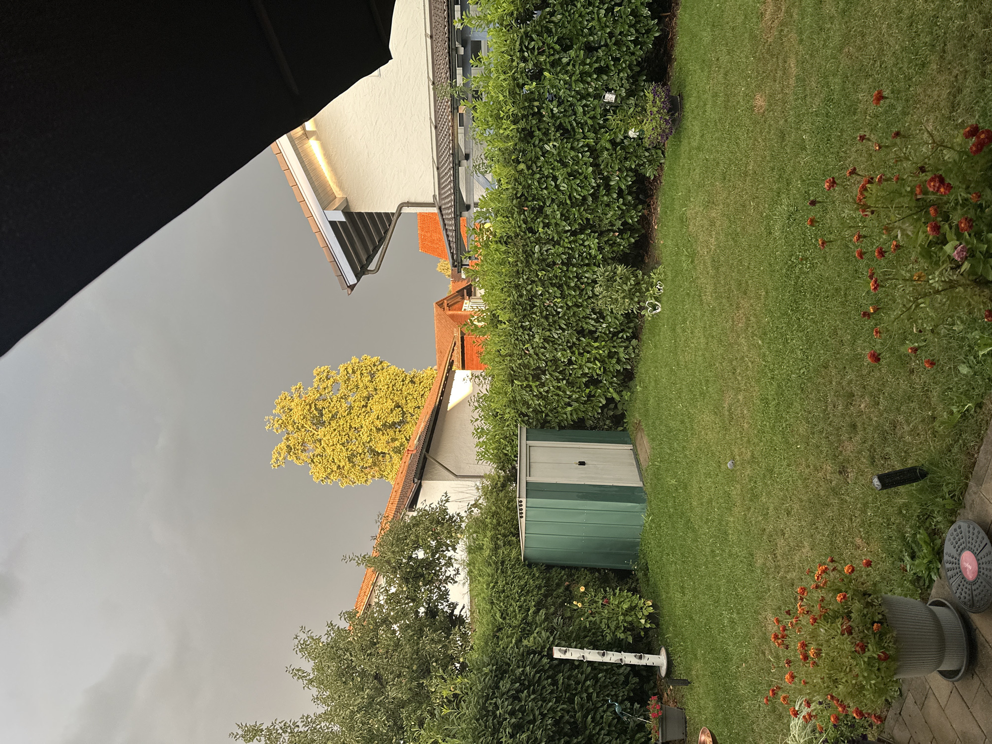 The image depicts a backyard in Bad Endorf, Germany, under a heavily overcast sky, suggesting impending rain. In the foreground, a well-maintained green lawn is visible, bordered by a dense, manicured hedge. To the left, a grey planter holds a cluster of vibrant orange flowers. Adjacent to the planter is a white, tiered garden ornament with small openings. A small, green garden shed with a grey door stands centrally within the yard, partially obscured by the hedge. In the background, residential buildings with tiled roofs are present, along with a tall tree displaying bright yellow foliage. A section of a dark awning extends from the top right of the frame, casting a shadow. The scene is devoid of human presence.