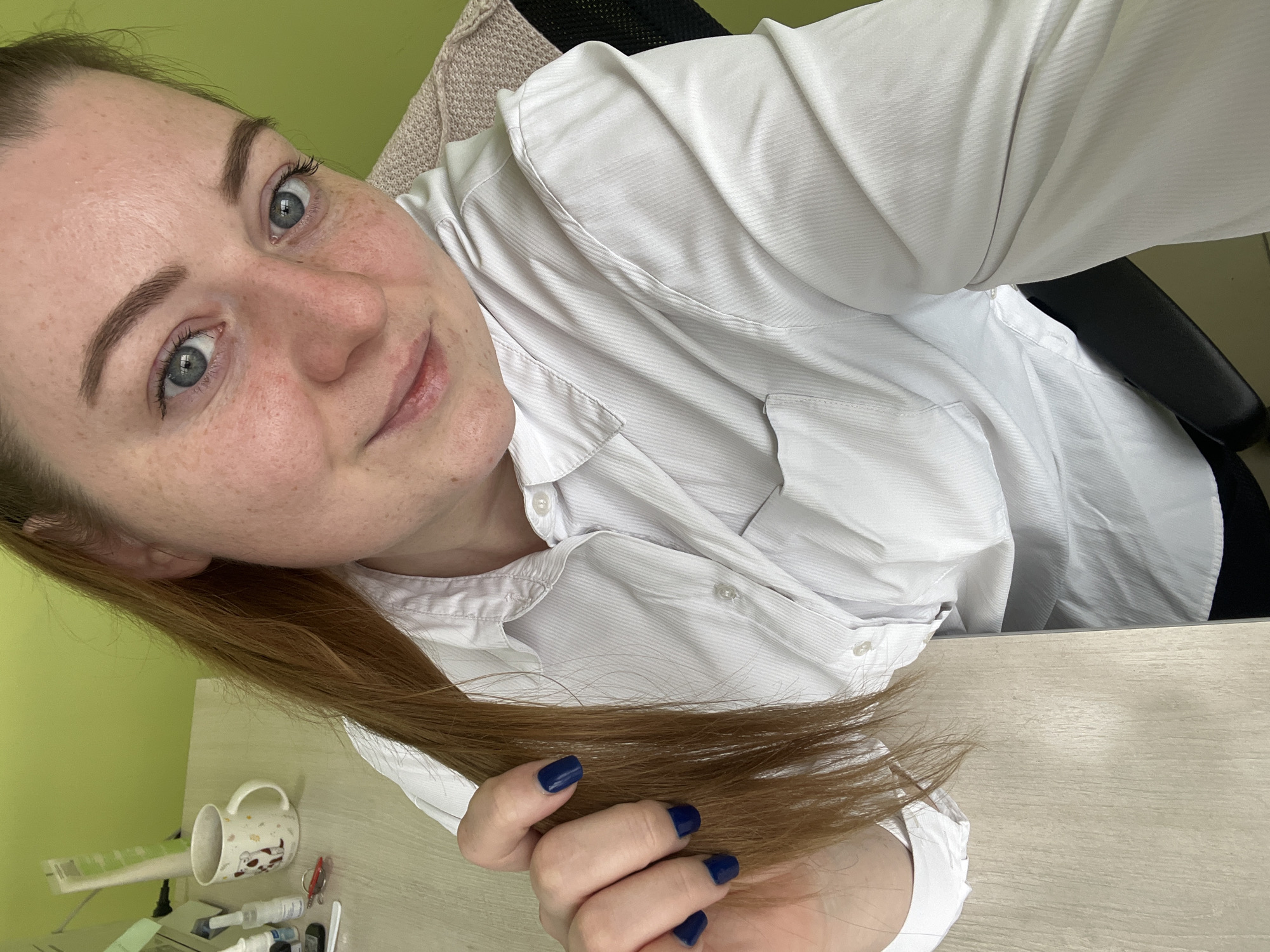 A close-up, tilted selfie captures a young woman in an indoor setting. She has fair skin with visible freckles, light blue-grey eyes, and medium brown hair with auburn undertones, which she holds a section of with her left hand. Her fingernails are painted a dark blue. She wears a white, button-up shirt with a subtle vertical pinstripe texture and a collar, appearing to be made of a textured fabric. Her expression is gentle, with a slight smile and a direct, relaxed gaze towards the camera.
The background features a plain light green wall behind her head. To her side and below, a light-colored wooden desk surface is visible, upon which various items are placed. These include a white mug adorned with a cartoon pattern (possibly cows or pigs), and several small, indistinct objects like tubes and clips, suggesting a workspace or personal area. Part of a chair, with a black frame and a beige fabric backrest, can be seen behind her. The lighting is bright and even, indicating daytime. The location context provided for the image is Klin, Russia. No visible text is clearly legible on any objects in the frame.