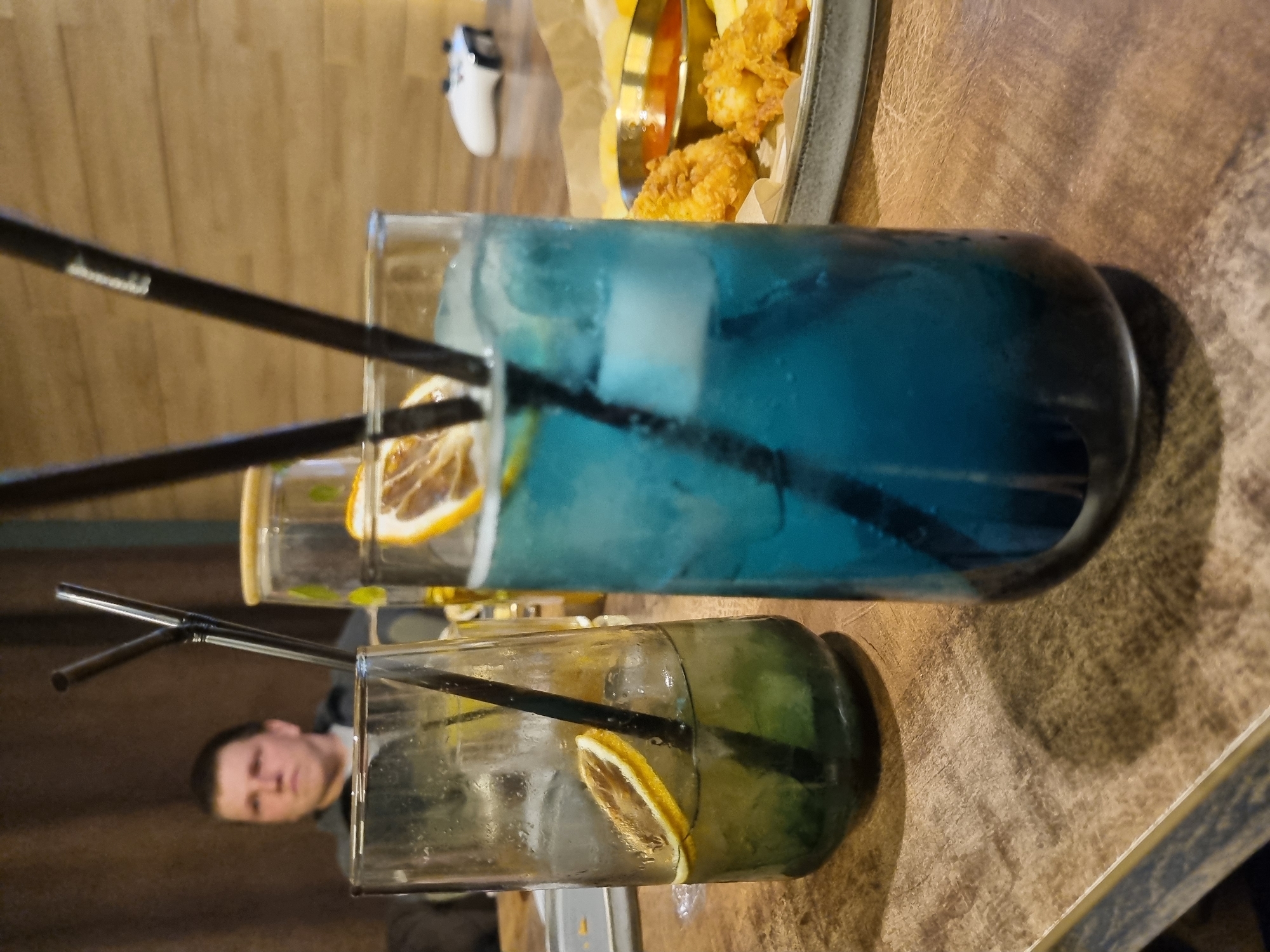 The image, taken in Chaikovsky, Russia, depicts a tilted view of a casual dining or bar setting with two drinks and food on a light-colored wooden table.
In the foreground, two tall, clear glass containers hold beverages. The larger drink, prominent on the right, is a bright blue color, filled with ice cubes, and garnished with a slice of dried orange. Two black straws are inserted into it. One of the straws bears faint, stylized text, which appears to be "Sturm" or a similar brand name. The second drink, positioned slightly to the left and closer to the bottom, is a lighter, greenish-yellow liquid, also with ice and a dried orange slice, containing one black straw.
Behind the drinks, a gray, textured plate holds a serving of golden-brown fried food, consisting of what looks like crispy chicken pieces or shrimp, alongside french fries. A small, shiny metal container with red dipping sauce is also on the plate.
In the upper background, to the left of the food, a white game controller, likely an Xbox controller, rests on a blurred light brown surface. A man's face, with short dark hair and a neutral expression, is partially visible in the mid-ground, appearing either reflected in the glass or through the contents of the smaller drink. The overall scene suggests a relaxed atmosphere, likely indoors, where people are enjoying refreshments and possibly entertainment.