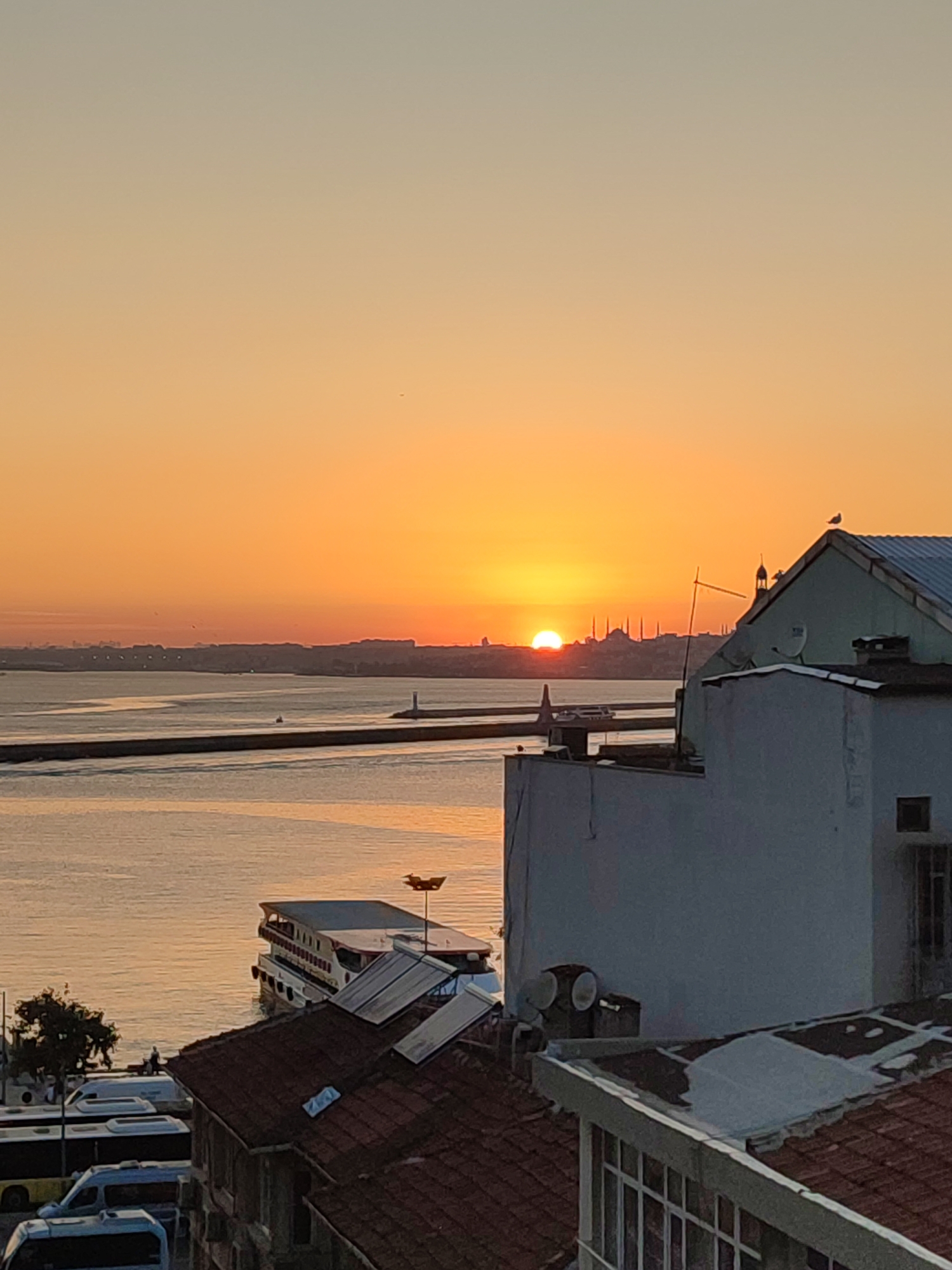 The image captures a sunset over the Bosphorus Strait in Kadıköy, Turkey. The sun is a bright orange orb, partially set, casting a warm glow across the sky and water. In the foreground, there are several rooftops with terracotta tiles, some with solar panels and satellite dishes. A white building with light blue trim stands prominently on the right. A large ferry boat is docked on the left side of the image, with several buses parked along the waterfront. In the distance, the silhouette of the Istanbul skyline is visible, featuring minarets and domes of historic mosques. A lighthouse stands at the end of a pier extending into the water. A single seagull is perched on the rooftop of the white building. The water appears calm, reflecting the colors of the sunset. The overall scene is one of serene urban landscape at dusk.