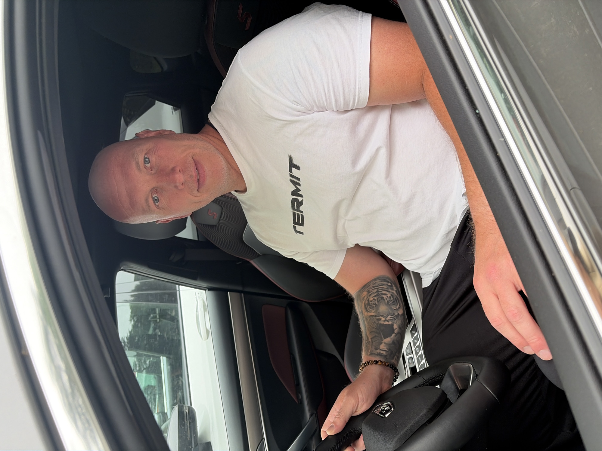 A bald, fair-skinned man in a white t-shirt with "TERMIT" printed on it is sitting in the driver's seat of a car. He is wearing black pants. His left arm is resting on the steering wheel and has a large tiger tattoo and a beaded bracelet. The car's interior is black and red. The man is looking at the camera with a slight smile. The scene appears to be in a parking lot, with other cars visible through the windows. The lighting suggests it is daytime. The city of Chaikovsky, Russia, is not evident in the image.