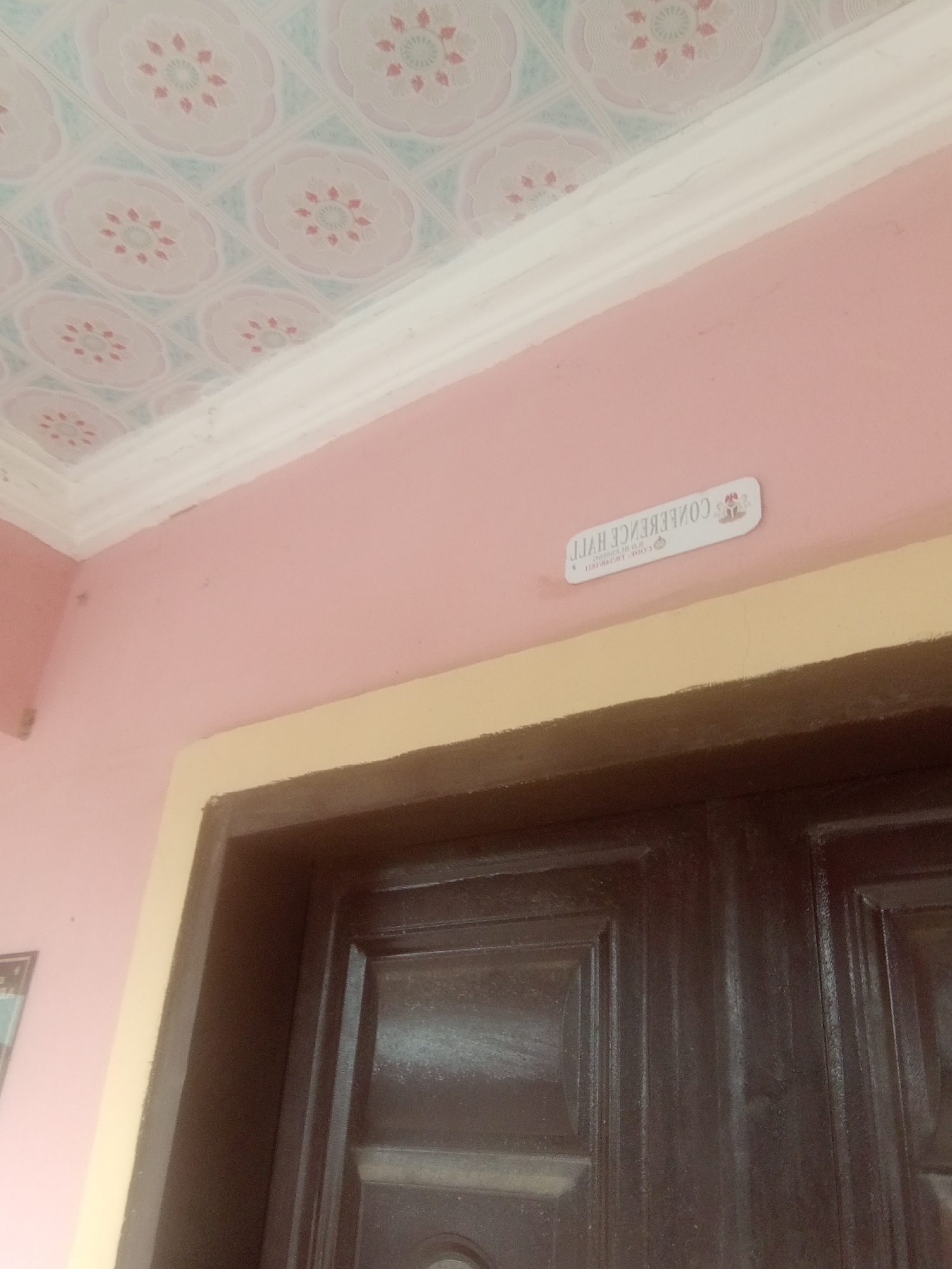 The image shows a ceiling with a floral pattern and a white crown molding. Below the molding, a pink wall is visible, with a light yellow trim above a dark brown door. A rectangular sticker is affixed to the pink wall, angled so that the text is upside down. The sticker has a white background and a thin gray border. At the top of the sticker, there is a small image with red and blue elements and what appears to be a coat of arms or emblem. Below this, text is printed in black. The text reads "LLAH ENEKEBNOC" which is "CONFERENCE HALL" reversed. Below this, in smaller text, also reversed, is "TIRIHIR TIO TI". There are no people visible in the image. The setting appears to be indoors, possibly in a hallway or entrance area, given the presence of a door. The lighting suggests it is daytime, but there are no cues about the weather. The sticker indicates that this is a conference hall. The location context provided is Jalingo, Nigeria.
