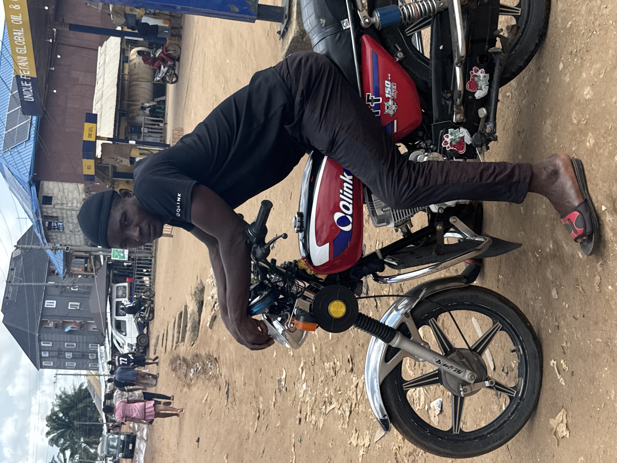 This daytime image portrays a vibrant street scene in Ihiagwa, Nigeria. In the foreground, a man with dark skin, wearing a black t-shirt, black pants, and a black cap, leans over a red motorcycle, looking directly at the camera with a neutral expression. He wears red and black sandals. The man's t-shirt features "QOLINK" text on the sleeve.
The motorcycle is a central subject, predominantly red with white and blue decals. Its tank clearly displays "Qlink," while the side fairing reads "150.. TIGER." Small "LOVE" stickers, resembling fruit, are affixed to the motorcycle's front forks, which also bear the word "SUPER." The front wheel has black spokes with silver details.
The background unfolds a typical urban or semi-urban landscape. To the left, a blue-roofed structure with solar panels is visible, beneath which stands a fuel station. A prominent yellow sign above reads "UNIQUE PETANI GLOBAL OIL &." Fuel pumps are discernible, with one clearly marked "DIESEL" and another "EZ." The ground is an unpaved mix of dirt and sand, strewn with some debris. In the mid-ground, several pedestrians are seen walking, and parked vehicles, including a white van and other motorcycles, line the street. Various buildings, some with corrugated roofs, and a multi-story structure contribute to the town's appearance. Palm trees are visible in the distance under an overcast sky, suggesting daylight.