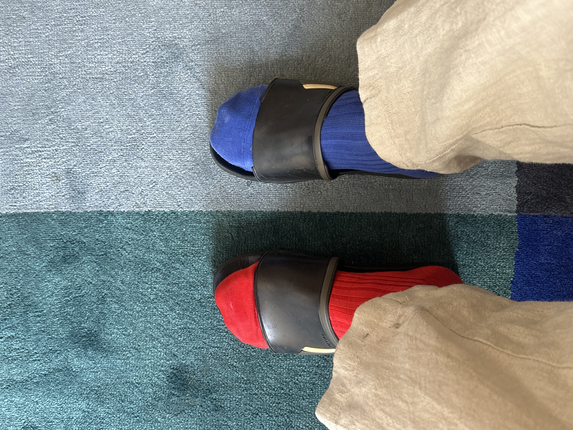 The image shows a top-down view of two feet wearing socks and sandals. The left foot has on a red sock and a black sandal, and the right foot is wearing a blue sock and a black sandal. The person is wearing tan-colored pants. The feet are positioned on a carpet with blue and green stripes. The exact location in London, United Kingdom cannot be determined from the image.