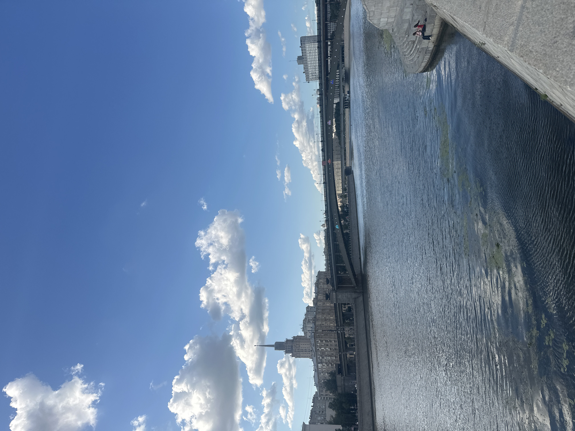 This is an image of the Moskva River in Moscow, Russia, on a sunny day with scattered clouds. In the foreground, the river occupies the majority of the frame, with ripples on its surface and patches of green algae. A curved concrete embankment lines the right side of the river, with two people, a woman and a girl, sitting on the steps. Across the river, a wide bridge with multiple levels spans the water, supporting traffic. Further back, buildings line the riverbank, including a prominent Stalinist skyscraper with a spire. The sky is a clear blue, with fluffy white clouds.