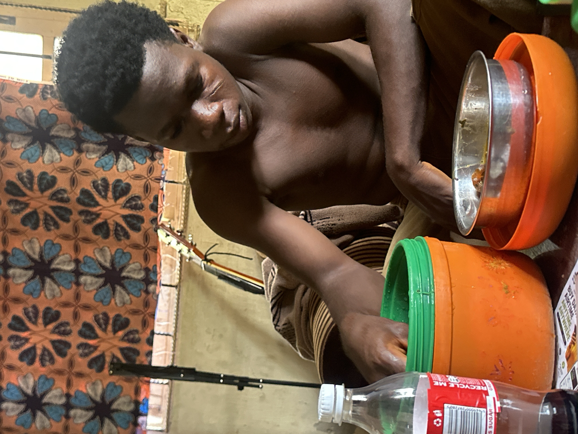 In Mararaba, Nigeria, a young, dark-skinned male with short, kinky black hair and prominent facial scars on his left cheek is seated indoors, shirtless, with his torso facing slightly left. He is actively reaching his right hand into a large, orange plastic bucket with a green lid. To his immediate left is a clear plastic bottle with a white cap, displaying a red label with a barcode and text including "RECYCLE ME" and "LESS SUGAR." To his right, a small silver metal bowl containing food rests inside a larger, orange plastic bowl. The background features a patterned textile hanging on the wall, exhibiting a repeating motif of dark and light blue heart-shaped petals arranged in a floral pattern on an orange-brown background. Visible in the background are parts of a black tripod stand and the neck and tuning pegs of an acoustic guitar. The individual is also partially covered by a brown patterned blanket or garment draped around his lower body.