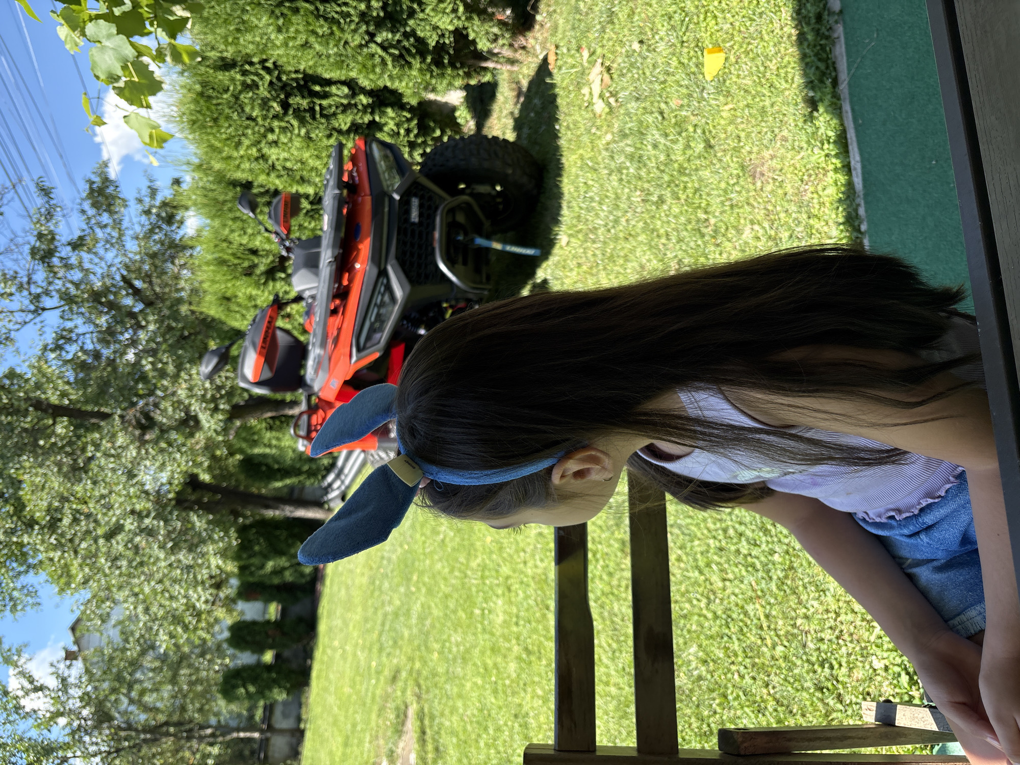 An outdoor scene in Oreshak, Bulgaria, captures a young girl from behind, seated on a wooden bench, facing away from the camera. She has long, dark hair and wears blue bunny ears on her head. Her attire consists of a light-colored tank top and denim shorts. In the background, a bright orange ATV is parked on a lush green lawn, surrounded by manicured evergreen bushes and mature trees. The sky above is a vibrant blue with scattered white clouds. A white building is visible in the distance, partially obscured by trees. The overall atmosphere suggests a sunny day in a residential or recreational outdoor setting.
