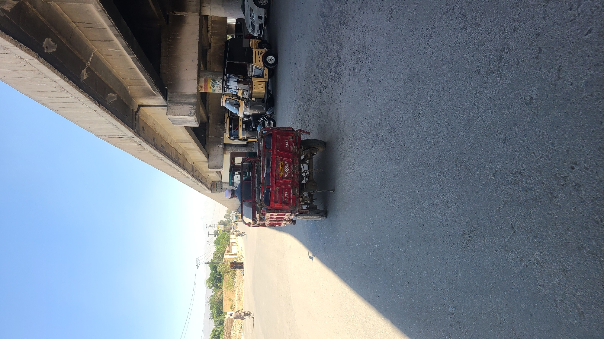 The image shows a street scene in Quetta City Tehsil, Pakistan, with a concrete overpass structure dominating the left side of the frame. Several auto-rickshaws (tuk-tuks) are parked under the overpass, including a red one in the foreground. The road surface is gray and appears slightly worn. In the background, across the road, there is a glimpse of trees, power lines, and some buildings. The sky is clear and blue. The lighting indicates it's daytime.