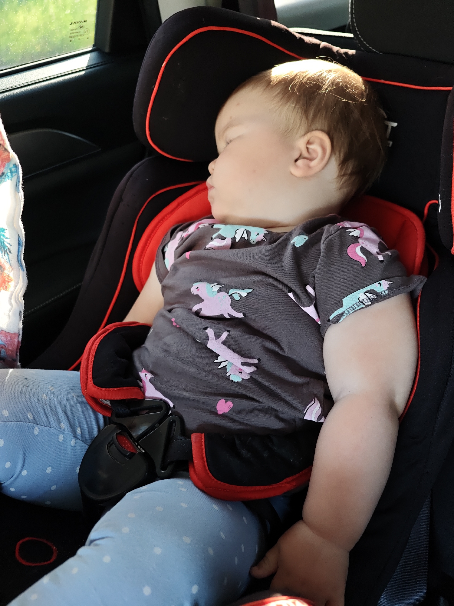 A baby sleeps in a car seat. The baby is wearing a gray shirt with unicorns and light blue polka-dotted leggings. The car seat is black and red. The setting is inside a car. There is no indication of the location being in Chaikovsky, Russia.