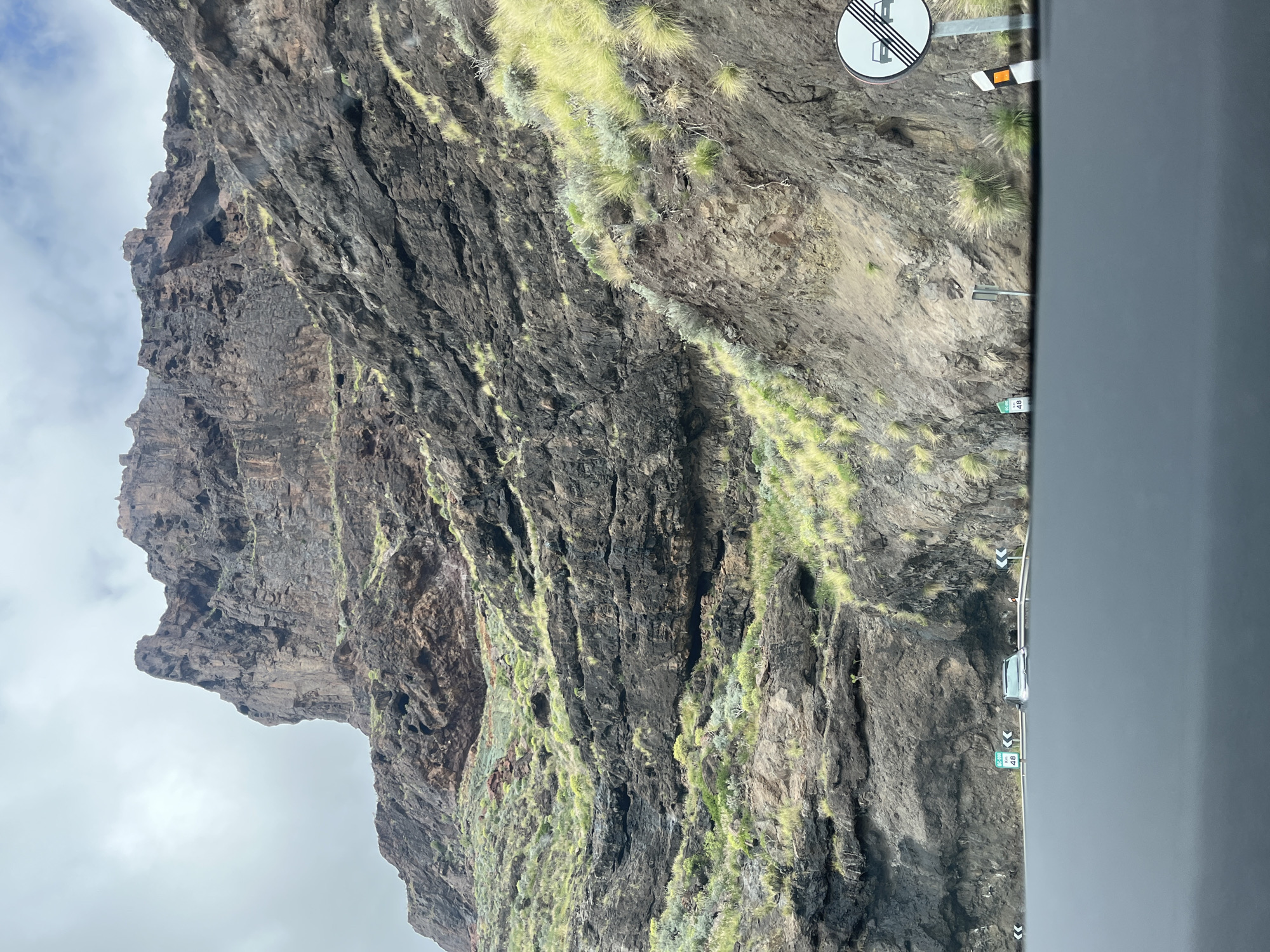 The image shows a steep, rocky mountain face covered with some green vegetation, likely taken from a moving vehicle on a road alongside. A road sign is visible, suggesting a winding road. The location is likely Mogán, Spain, based on the rugged landscape and potential for coastal mountain roads. A kilometer marker "48" can be seen next to the road. The weather appears overcast.