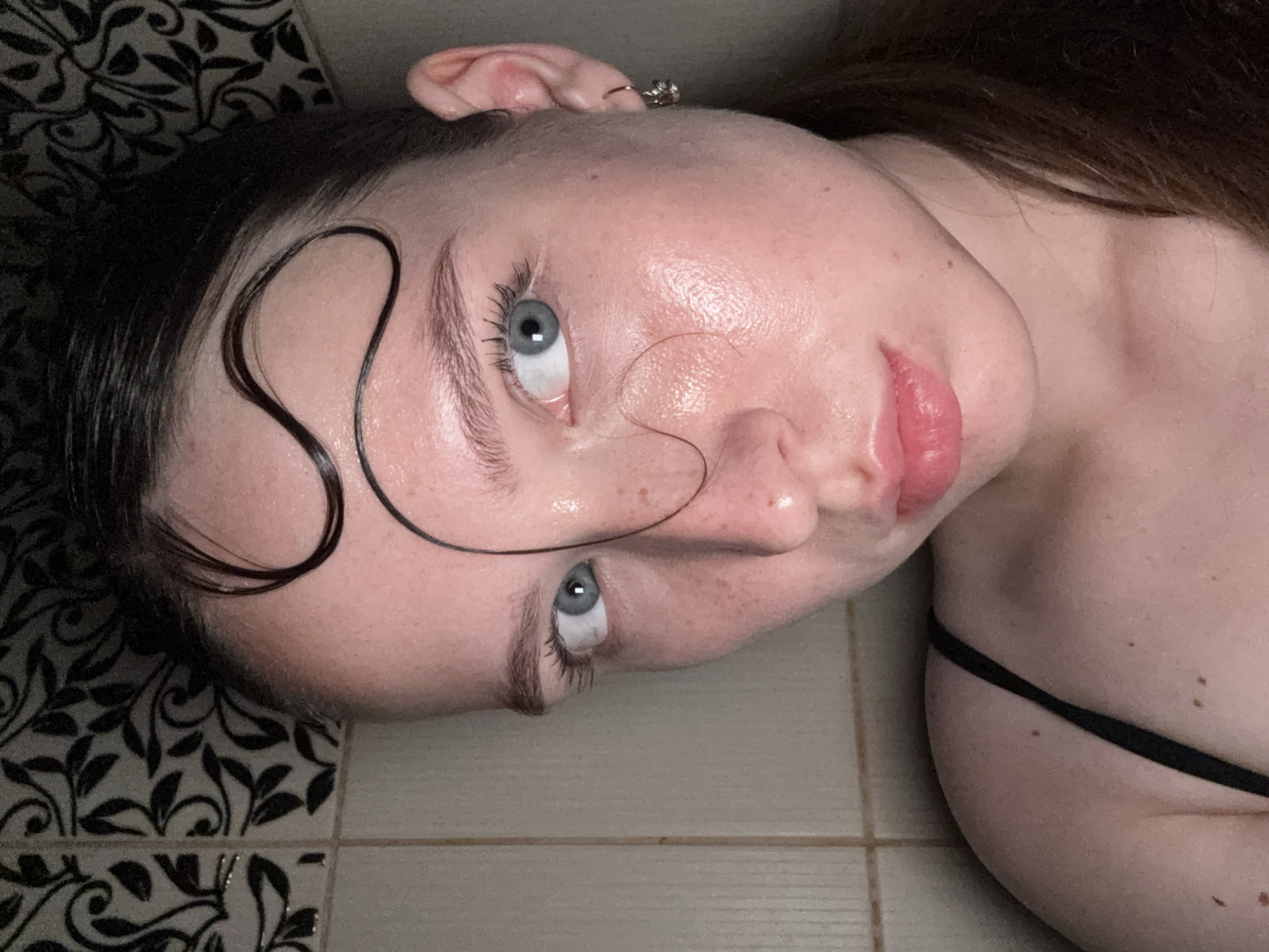 A close-up portrait, taken in Chaikovsky, Russia, depicts a single female individual facing the viewer. The subject has fair skin, prominent blue eyes, and dark brown hair, which is pulled back, with several wet, dark strands artfully shaped into curves and S-patterns across her forehead. Visible freckles are present on her cheeks and forehead. Her skin exhibits a hydrated, slightly glossy sheen. Her lips are full, natural pink, and slightly parted. She is wearing a black, thin-strapped garment, and a small silver earring is visible in her right ear. The background consists of a tiled wall, primarily beige with dark grout lines. A section of the wall in the upper left displays a light-colored tile with a dark, intricate floral or leafy pattern. Lighting is ambient, illuminating the subject's face directly.
