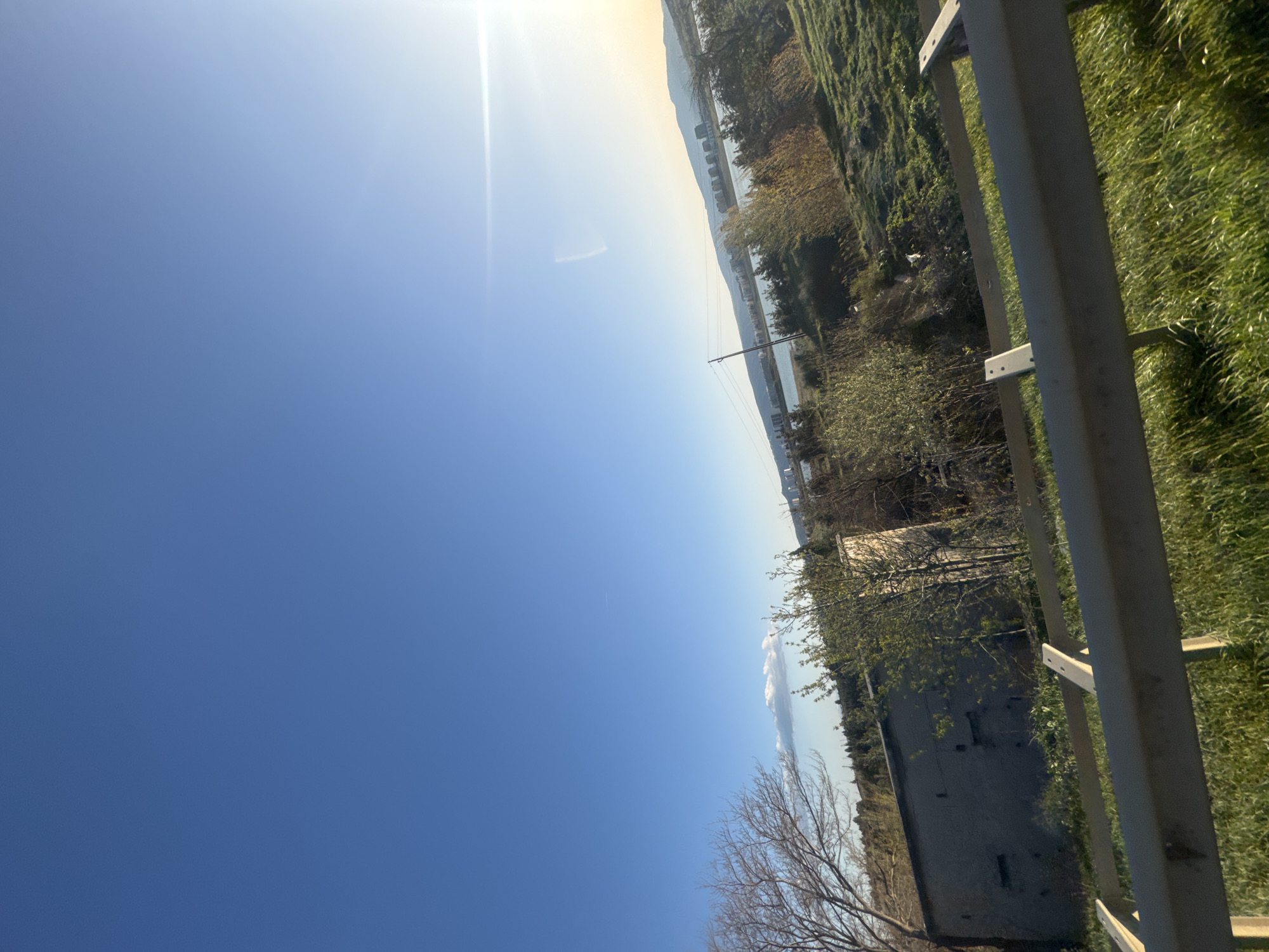 The image shows a landscape scene under a clear, bright sky. The sky occupies a large portion of the frame, transitioning from deep blue to lighter hues near the horizon where the sun shines. Along the horizon, there is land with greenery, trees, and buildings, possibly including residential or commercial structures. A body of water is faintly visible, separating the foreground from distant mountain ranges. A fence runs along the right side of the image.
It is not possible to determine the specific location of Tbilisi, Georgia, from the provided image.