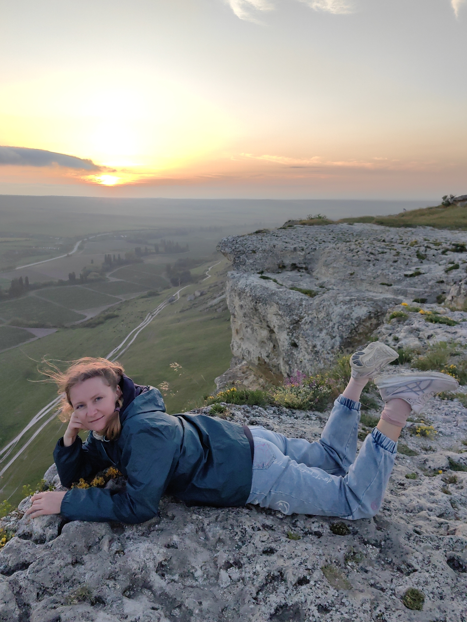 A woman is lying on a rocky cliff, overlooking a vista during a sunset. She is wearing a dark green hooded jacket and light blue jeans with elastic cuffs. Her legs are raised slightly, and she has on light-colored athletic shoes and pink socks. Her head is resting on one hand.
The location is unknown. The background features a landscape with fields and a road leading into the distance, under a sky with a prominent sunset.