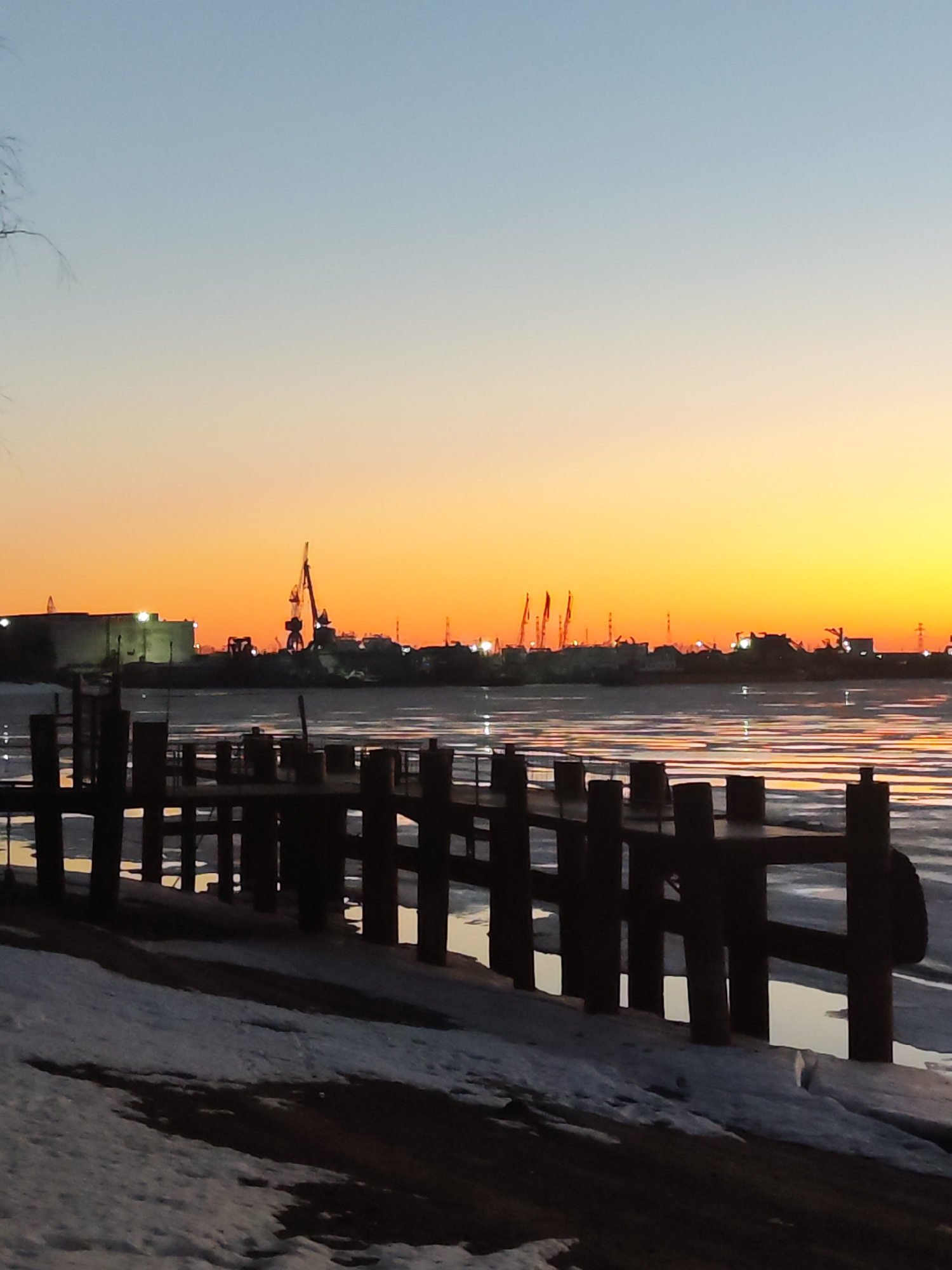 The photo captures a twilight scene in Chaikovsky, Russia, featuring a landscape with a mix of natural and industrial elements. A wooden pier stands in the foreground, with snow on the ground next to it. Beyond the pier, the river is partially frozen with reflections of the twilight sky visible on the water surface.
In the background, the cityscape is silhouetted against the colorful sky, which transitions from a pale blue at the top to a bright orange near the horizon. Industrial structures, including cranes, are visible along the skyline, suggesting a port or industrial area. The scene is lit by the twilight sky and reflections from the city lights.