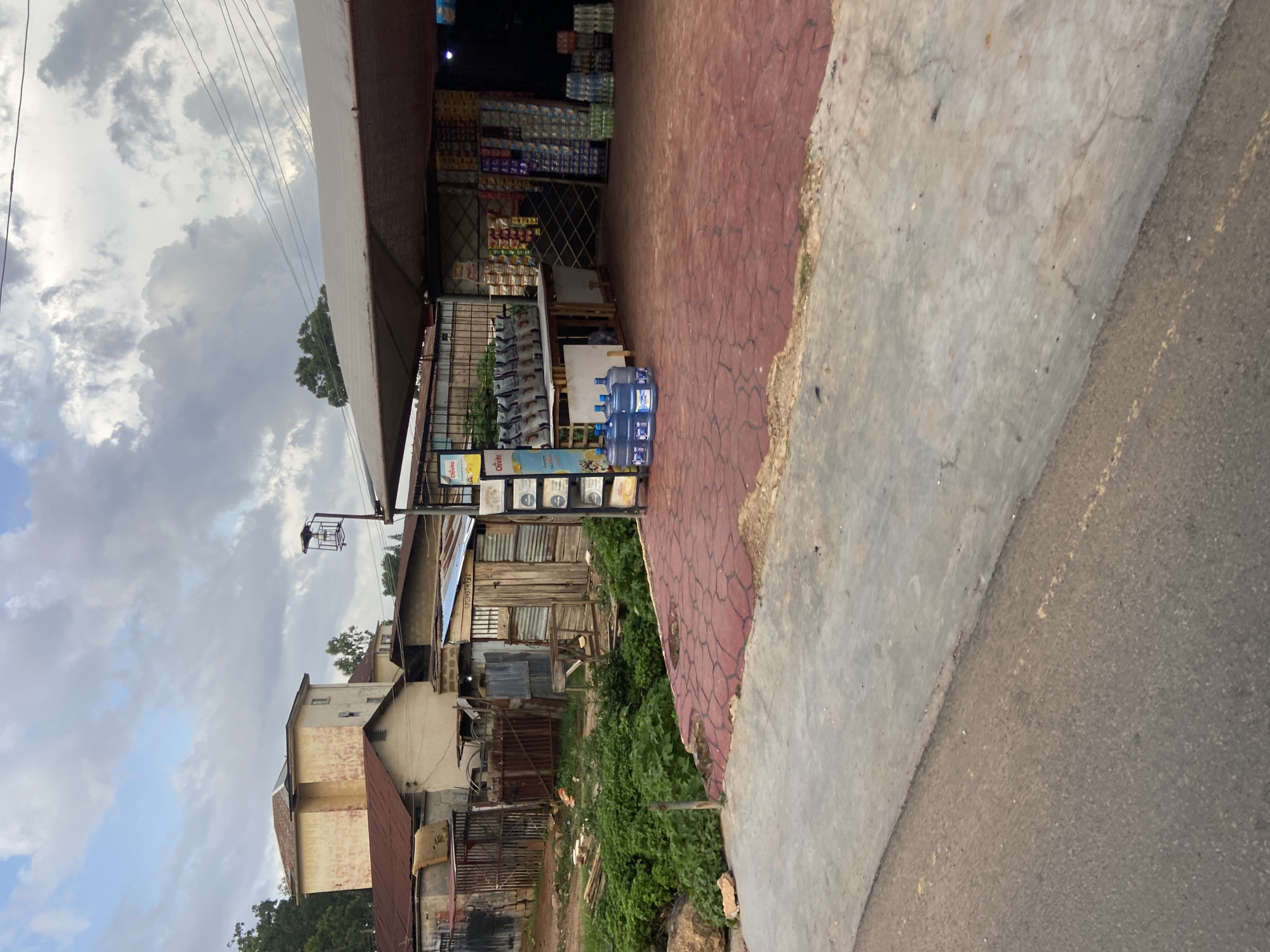 The image depicts a street scene in Jos, Nigeria, likely during the day under an overcast sky. The foreground is dominated by a paved area with red interlocking bricks, suggesting a walkway or part of a road. To the right, the paving transitions to a darker asphalt surface, indicating a street.
The main subject is a storefront, a small shop or market stall, set back from the paved area. The shop appears to be selling various goods, including stacks of bottled water, likely for sale. The interior of the shop is visible and stocked with shelves full of packaged items, possibly food or household goods. A sign above the shop entrance is partially visible, with some text that is difficult to decipher but appears to be in English.
Adjacent to the shop, there are structures made of corrugated metal and wood, indicating a less developed area. Green vegetation, possibly overgrown grass and bushes, grows in front of these structures, bordering the shop. In the background, buildings with a more permanent construction are visible, suggesting a mixed urban environment. The sky is cloudy, with a mix of light and dark gray clouds, giving the impression of a potentially rainy or humid day. There are no people clearly visible in the image, and the scene appears to be a quiet moment in a commercial area. The overall impression is one of a bustling, yet somewhat unkempt, local marketplace.