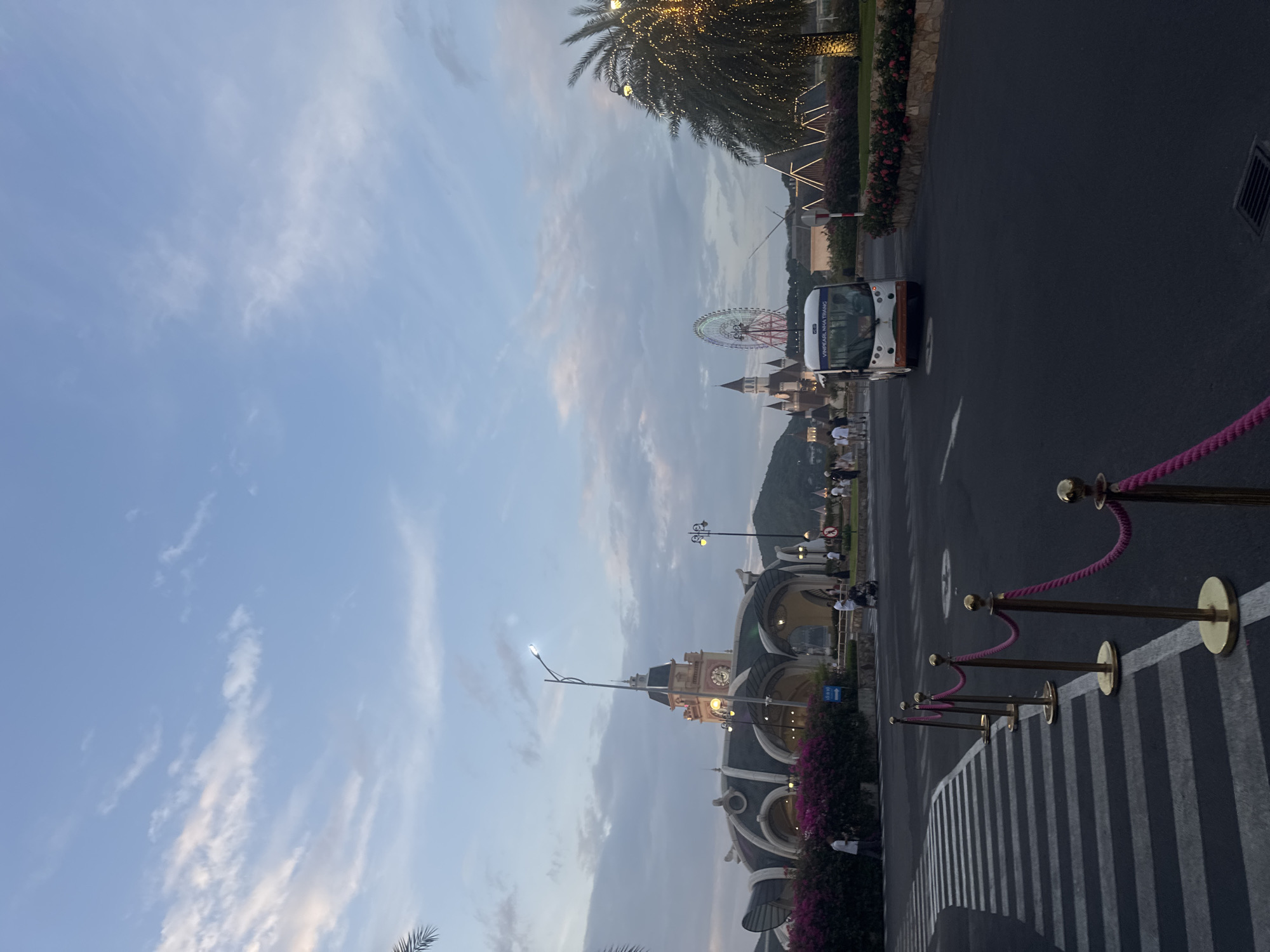 This is a photograph taken in the evening at Vinpearl Nha Trang, Vietnam. The image captures a view of the entrance area of the amusement park. In the foreground, a crosswalk marked with white stripes is visible, bordered by a line of stanchions with pink ropes. A dark asphalt road stretches into the distance, with a white lane marking.
Further into the scene, a white shuttle bus with the text "VINPEARL NHA TRANG" on its front is driving on the road. To the left of the bus, a large, ornate building with a clock tower is prominent, adorned with arched entrances and dark-tiled roofs. In front of this building, a dense hedge of purple flowers lines the pathway.
In the middle ground, several people are seen walking, some are pushing strollers, and others appear to be employees directing traffic. To the right of the bus, a large Ferris wheel, illuminated with colorful lights, stands against the dusky sky. Adjacent to the Ferris wheel, a palm tree decorated with strings of white lights is illuminated. Further back, a cluster of buildings with pointed roofs, resembling a castle, are visible. The sky above is a blend of blue and gray clouds, with the lighting suggesting it is either sunrise or sunset. Hills are visible in the far background.