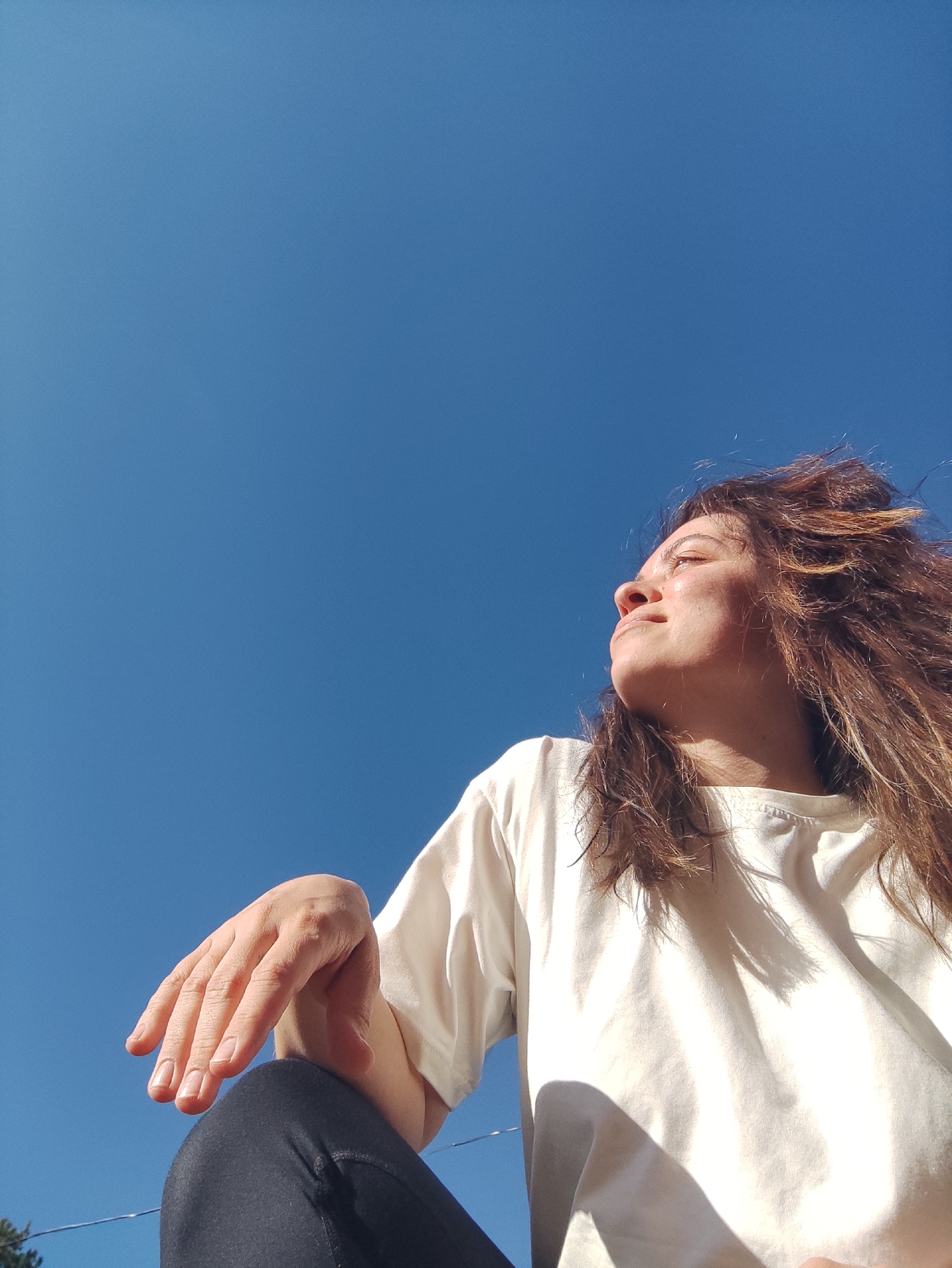 A low-angle outdoor shot captures a young woman looking up and to the right against a vast, clear blue sky. She has long, wavy, reddish-brown hair, which appears to be slightly wind-swept, framing her face. Her profile is illuminated by bright sunlight, causing her eyes to squint slightly and highlighting the texture of her skin and hair. She wears a plain white t-shirt, with shadows from the strong light falling across its fabric. Her right hand, with neatly trimmed nails, rests gently on her bent knee, which is covered by dark, possibly black, clothing. The background is almost entirely a brilliant, cloudless azure sky, suggesting a bright and sunny day. A tiny glimpse of green foliage and thin, dark lines, possibly wires, can be seen at the very bottom left corner of the frame. The woman's expression is serene and contemplative, as if she is enjoying the sun or observing something in the distance. The setting for this image is Фоки, Russia. No text is visible in the media file.
