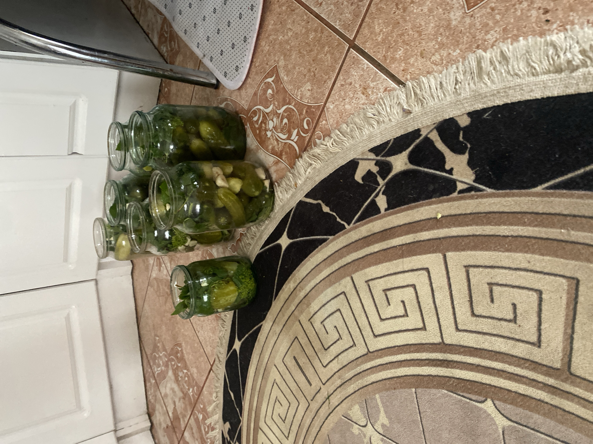 The image shows several glass jars filled with pickles, possibly on a kitchen floor. The jars are of various sizes and appear to be homemade. They are arranged on a tiled floor, with a portion of a round rug in the foreground. The background shows white cabinets. There are no people or animals visible in the image. The location of Ispas, Ukraine is not discernible from the visual information in the image.