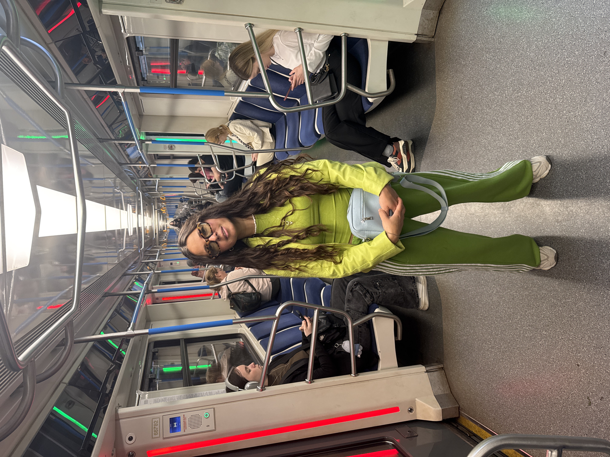 The image depicts the interior of a subway car in Kazan, Russia. A young woman with long, wavy brown hair and sunglasses is standing in the aisle, facing the camera. She is wearing a bright green tracksuit and holding a light blue purse. Other passengers are seated along the sides of the subway car. Some are looking at their phones, while others are simply sitting. The subway car has blue and white seating with metal frames. A horizontal red line is on the bottom of the seating. There are overhead handrails visible. The car is well-lit, and the floor is a speckled gray. The view down the length of the car shows more passengers.