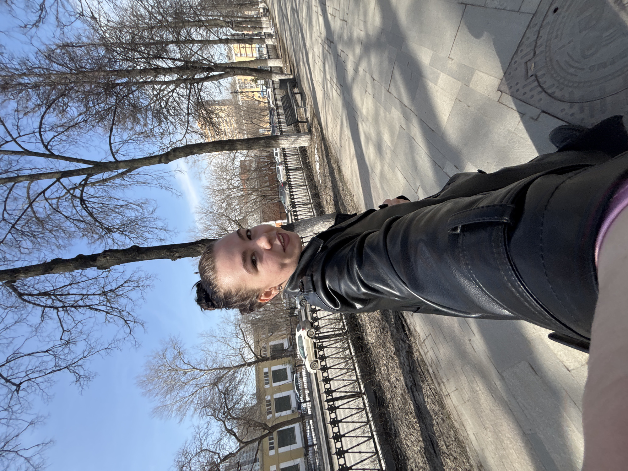 A person stands on a sidewalk in Perm, Russia. The sidewalk is paved with large, rectangular stones. A wrought-iron fence separates the sidewalk from a street where cars are parked. Bare trees line the sidewalk and the street. Buildings are visible across the street. The sky is blue. The person is wearing a black leather jacket. A manhole cover is on the sidewalk near the person.