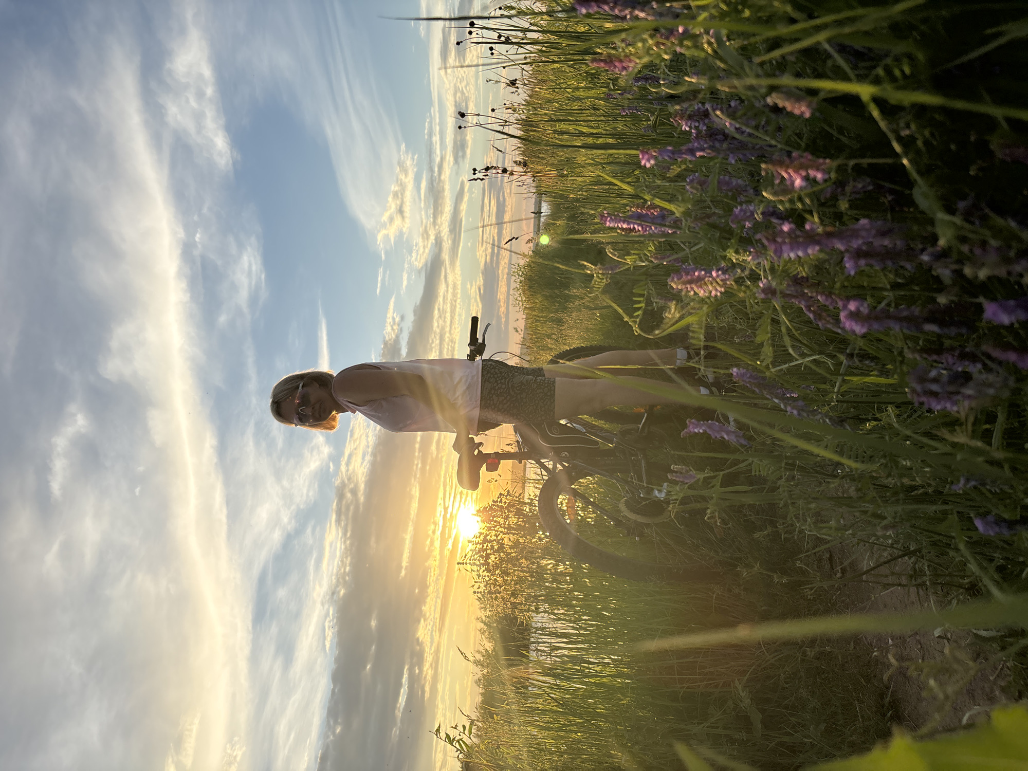 In this portrait, a person is captured on a bicycle at sunset. The individual is wearing sunglasses and a light-colored top, with patterned shorts. They are positioned on a dirt path surrounded by tall green grass and blooming purple flowers. The sky is a canvas of wispy white clouds, with the golden sun low on the horizon, casting a warm glow over the scene. The light reflects off the water in the distance, suggesting a lakeside or riverbank location. The overall mood is serene and highlights the beauty of nature during golden hour. The image was taken in Chaikovsky, Russia.