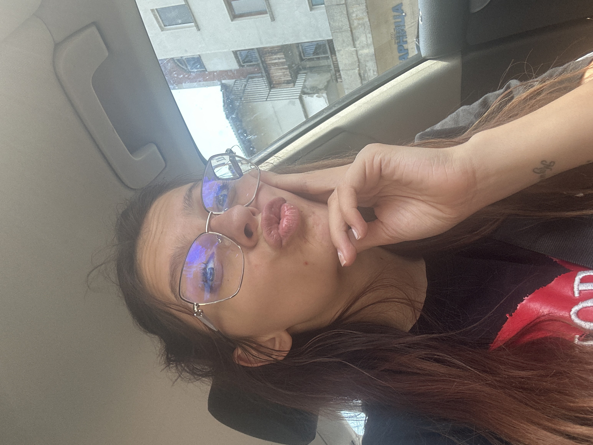 A young woman with long, dark brown hair and glasses is taking a selfie inside a car. She is wearing a black t-shirt with a red graphic design. Her right hand is resting on her chin, and her lips are pursed. The background shows buildings and a street scene, suggesting an urban environment. There is also a car interior visible, including a handle on the roof lining. The reflection in her glasses shows buildings and possibly a blue sky. The image appears to have been taken in Tryavna, Bulgaria, based on the text visible on a sign in the background which reads "АРПИЛЛА" and below it "AUDI". The overall lighting suggests daylight.