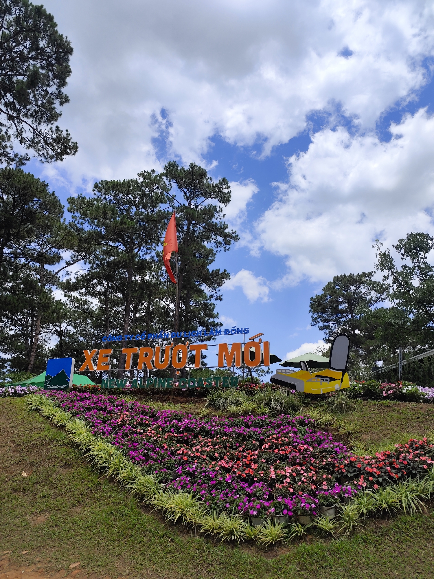 The image shows a floral display with a sign that reads "XE TRƯỢT MỚI" (which translates to "NEW ALPINE COASTER") in Da Lat, Vietnam. The sign is surrounded by colorful flowers arranged on a grassy hill. In the background, there are tall pine trees and a blue sky with scattered clouds. A Vietnamese flag is visible among the trees. A yellow vehicle model is placed alongside the sign. The floral arrangement is well-maintained and vibrant, suggesting a popular tourist location.