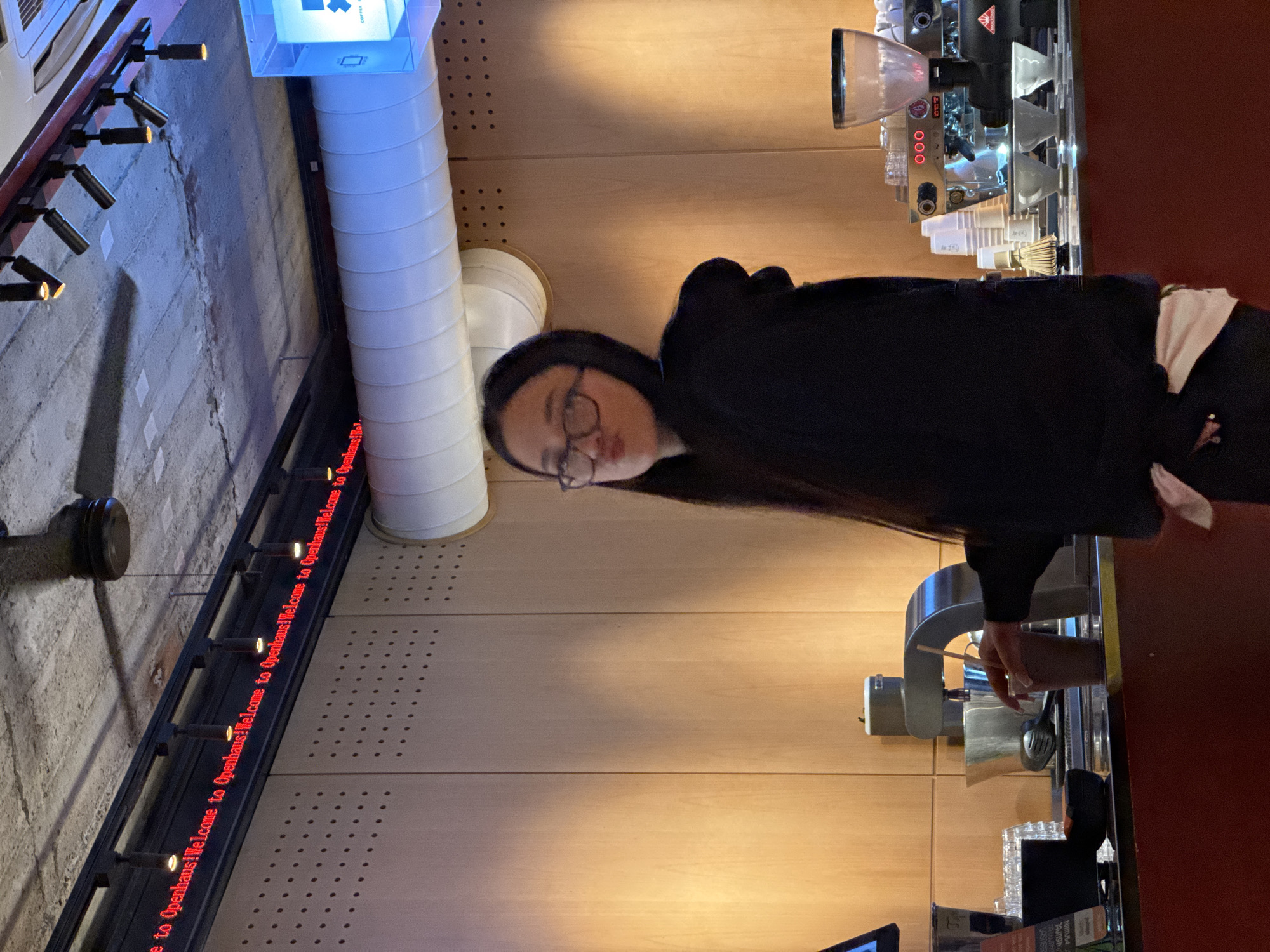 Here is a description of the image:
A vertically oriented image shows a person with dark hair and glasses standing behind a counter, appearing to make coffee using an espresso machine. The environment suggests a café or coffee shop with specific design elements: a large white ventilation tube is visible near the ceiling, and a red LED sign reads "Welcome to Openhaus!" The background includes a wall with a light wood paneling and a coffee-making machine. The location is Yerevan, Armenia, as suggested by the branding on products visible in the bottom right corner of the photo.