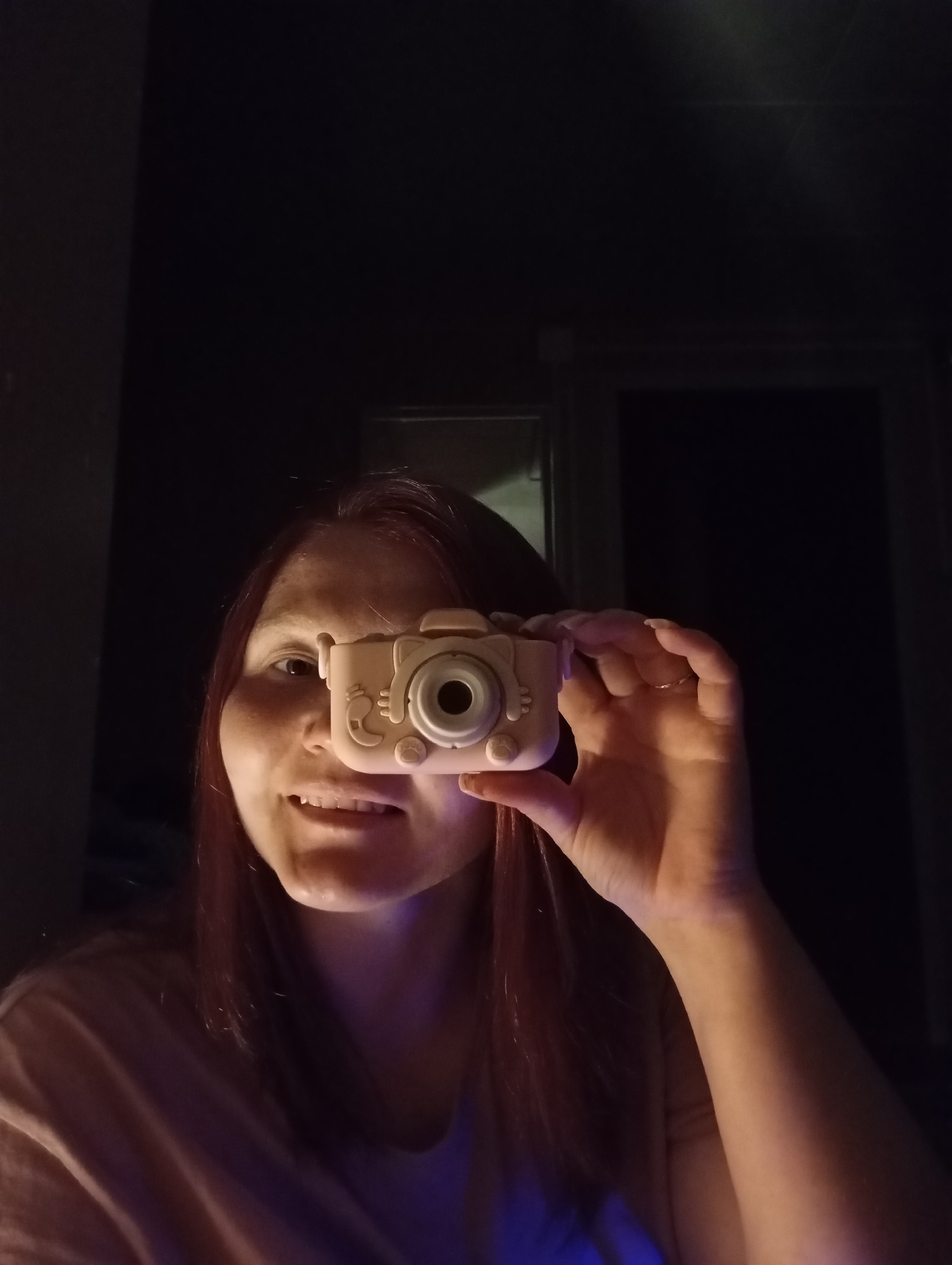 The image shows a woman indoors, holding a small, toy-like camera in front of one of her eyes. She is fair-skinned, with reddish-brown hair. She is wearing a light-colored t-shirt and smiling slightly. The background is dark, with a hint of a window or doorway visible behind her. The lighting appears to be from a source below, casting a blueish glow on her chin and neck. Location is Unknown, Unknown.