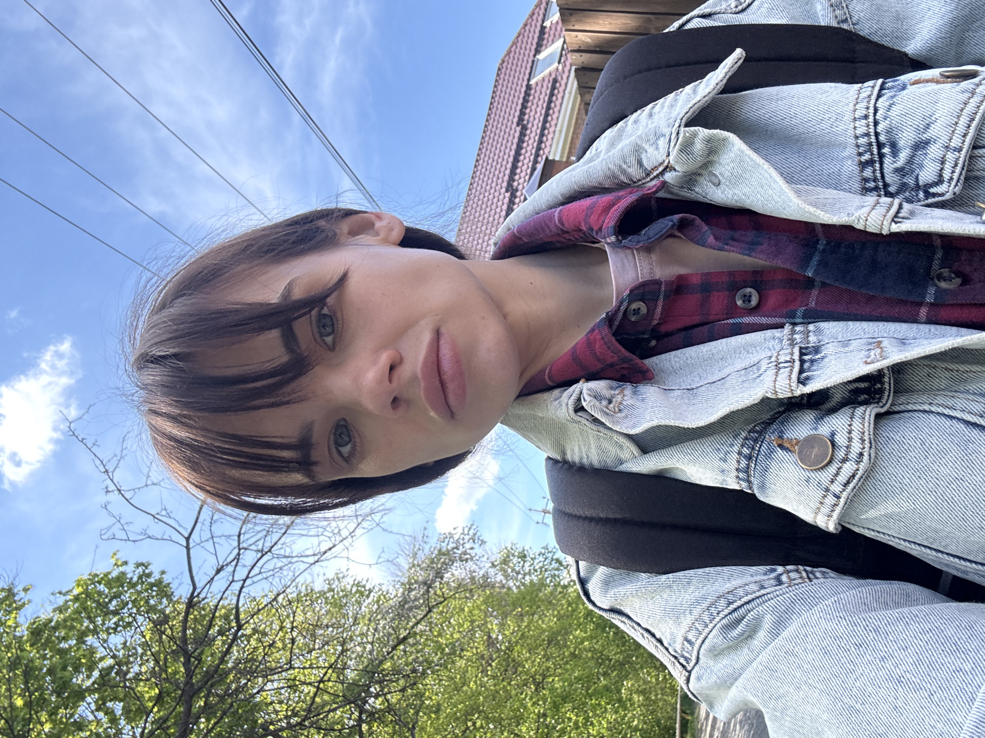 A woman with short brown hair and bangs, wearing a denim jacket over a red and black plaid shirt and a pink shirt, is standing outside on a sunny day. She has a black backpack on. Behind her, there's a blue sky with light clouds, trees with green leaves, and the corner of a red-roofed building. Power lines are visible against the sky. No information confirms this is in Rostov-on-Don, Russia.