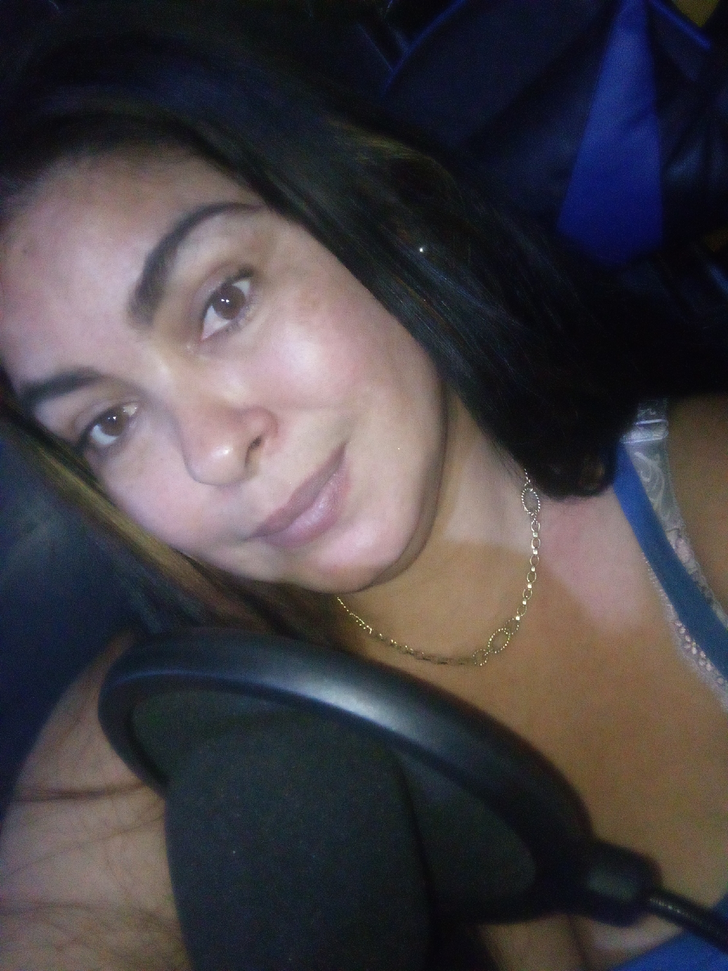 The image displays a close-up portrait of an adult female in an indoor setting, reportedly in Juan Griego, Venezuela. The individual has light-toned skin, dark brown hair, and brown eyes, gazing directly towards the camera with a neutral to slight smile. She wears a gold-colored chain necklace with oval links. A blue garment strap is visible on her right shoulder/chest area. A black microphone, likely studio equipment with a foam pop filter, is positioned in the lower-left foreground, partially obscuring her body. The background is dark and out of focus, revealing a dark blue object consistent with a chair back or furniture. The lighting appears to illuminate the subject's face from the front.