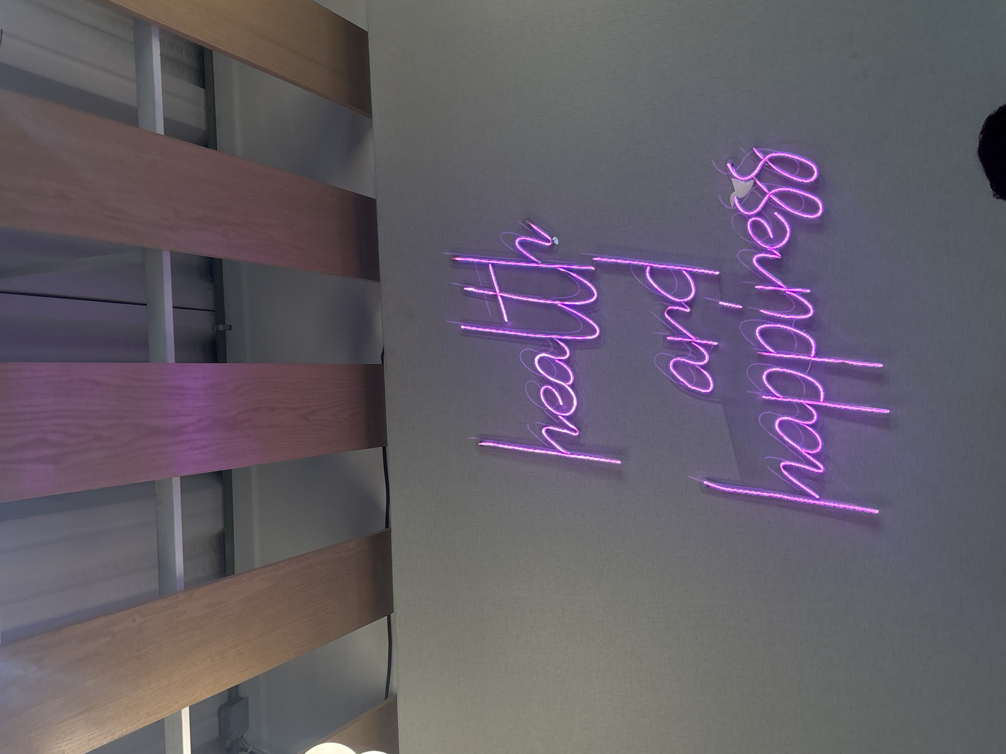 The image shows a portion of a wall displaying the neon words "health and happiness," and a part of a structure made of wood and metal. The location is not discernable in the image.